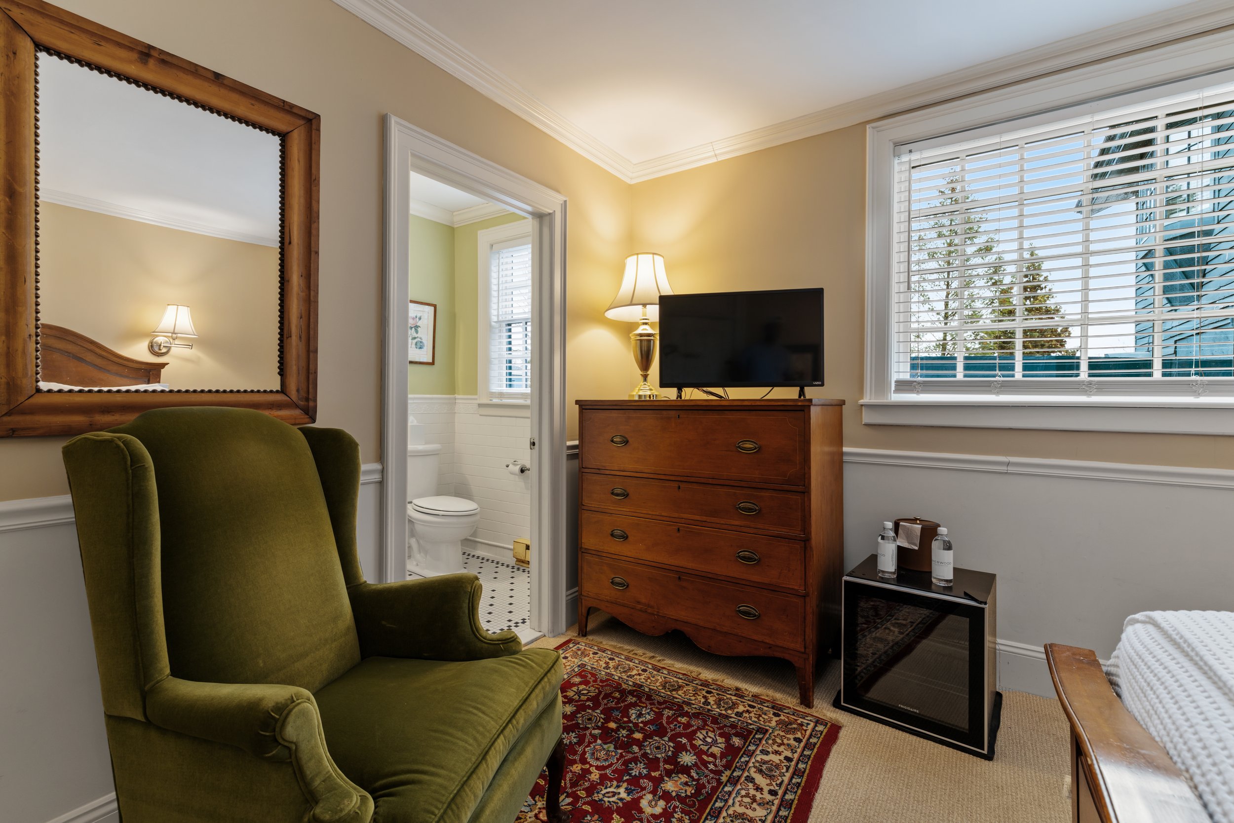 View of a cozy bedroom corner with a green upholstered armchair, a patterned rug, a wooden dresser with a TV on top, a bedside lamp, a window with white blinds, and a bathroom visible through an open door.