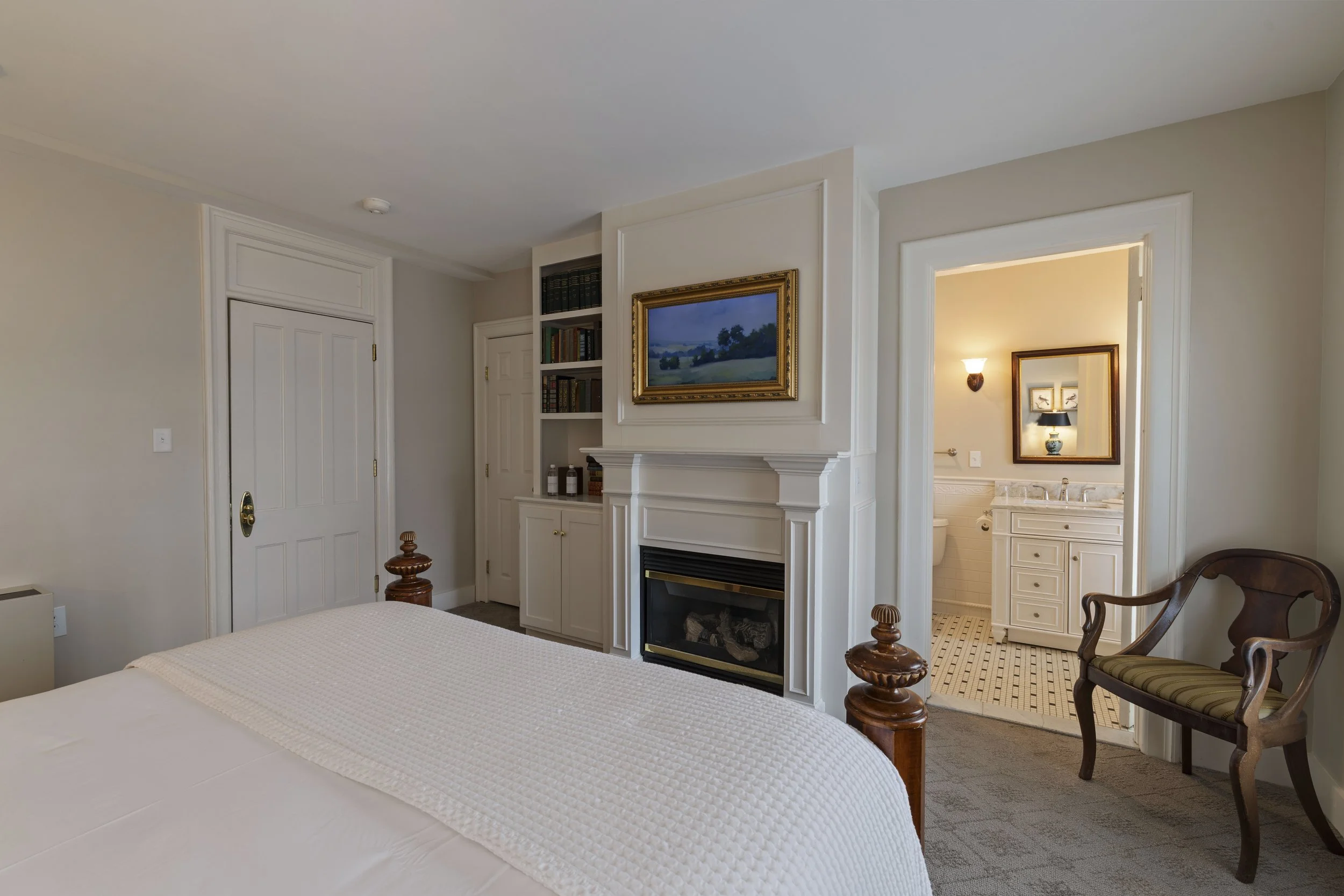 Hotel room with a bed, a fireplace, framed paintings, a built-in bookshelf, and a small bathroom visible through an open door.