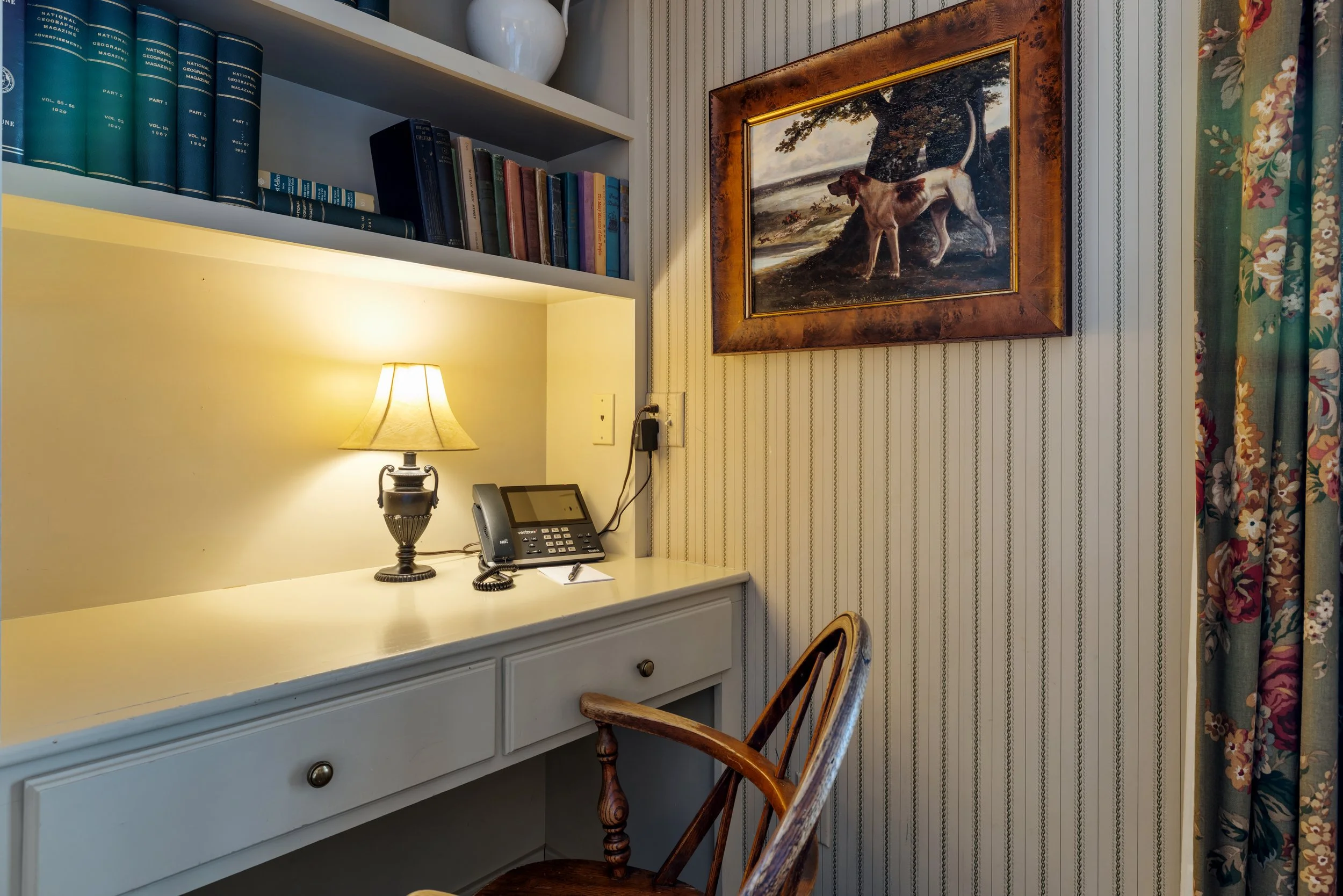 A cozy office nook with a white desk, a lamp, a telephone, and bookshelves above. The wall has striped wallpaper, and there is a framed picture of a dog in a natural setting. A wooden chair with curved arms is in front of the desk, and floral curtains are on the side.
