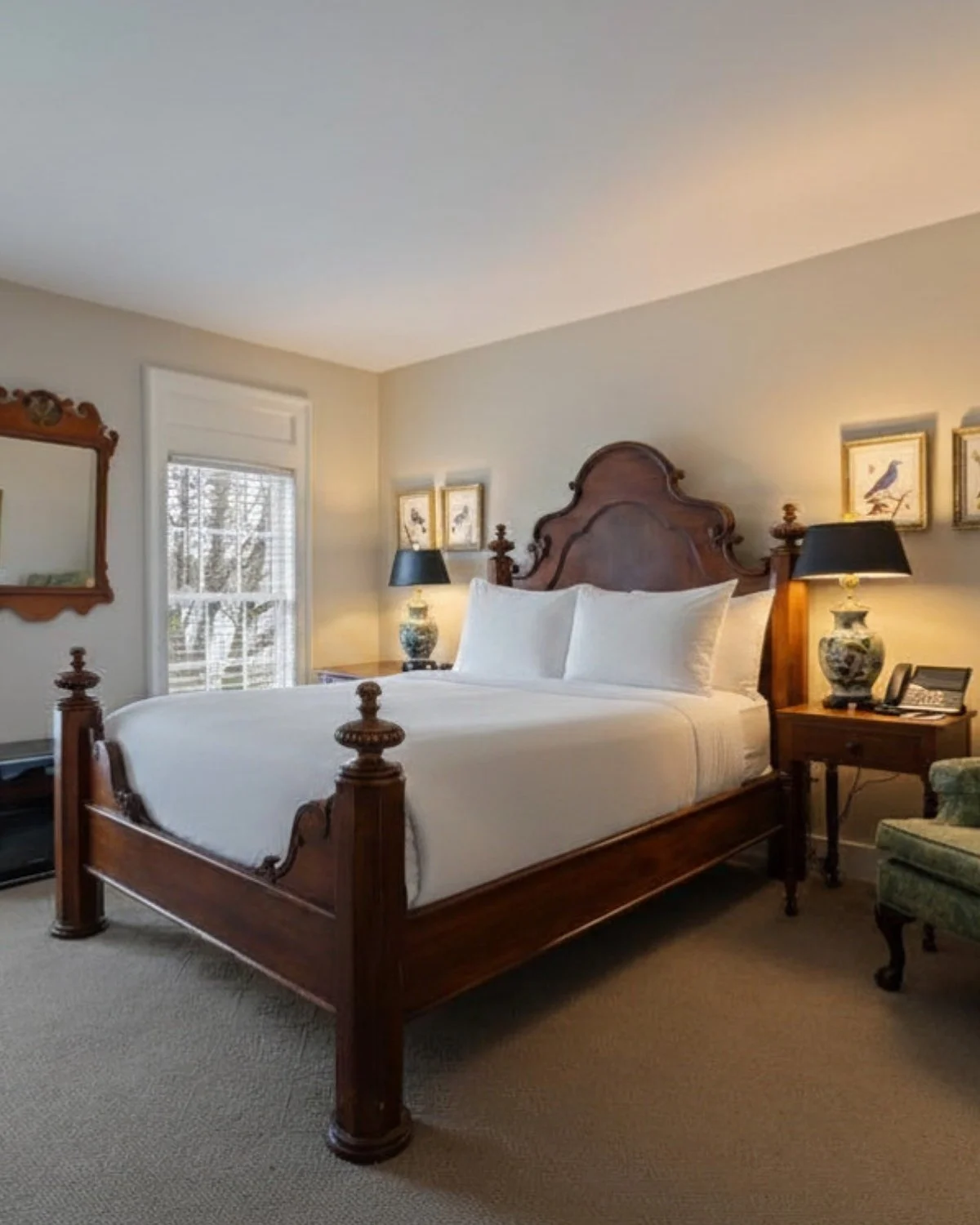 A bedroom with a wooden bed featuring a high headboard and posts, white bedding, two bedside tables with decorative lamps, a window with blinds, and framed bird artwork on the wall.