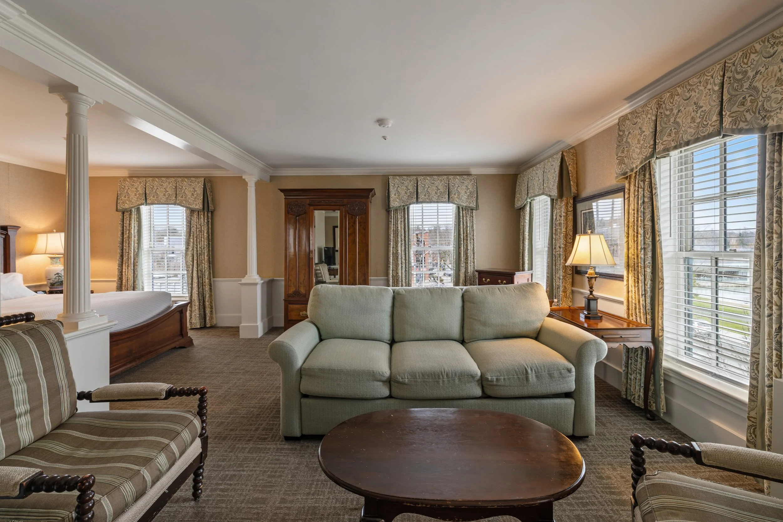 Hotel suite living room with large windows and beige curtains, a beige sofa, a wooden coffee table, a bed with white linens, lamps, and traditional wood furniture.