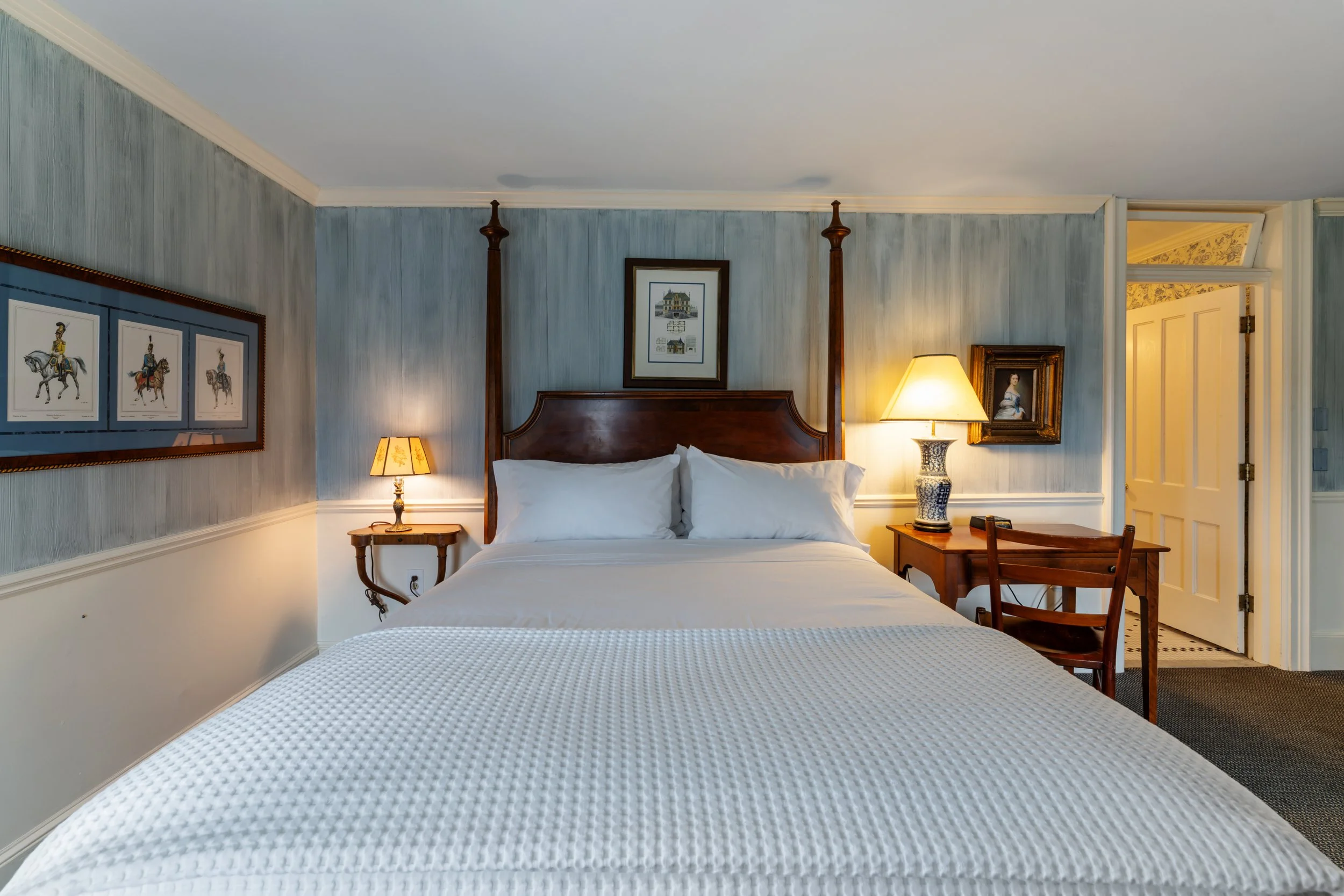 A cozy hotel bedroom with a white textured bedspread, wooden headboard, and matching bedside tables with lamps. The walls are decorated with framed artwork, including sketches of castles and a portrait of a woman. There's an open doorway revealing a room with patterned wallpaper.