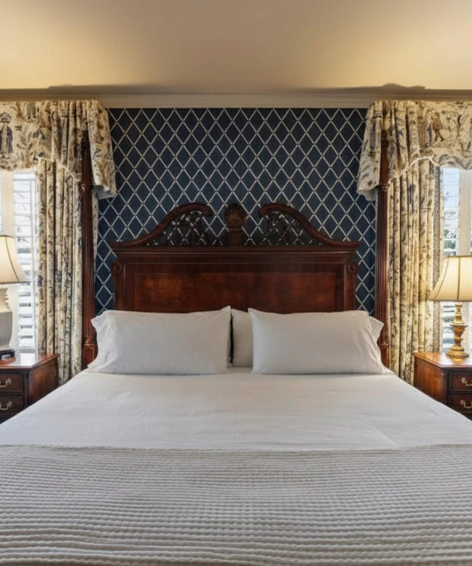 A hotel room with a king-sized bed, white bedding, two pillows, a wooden headboard, dark blue patterned wallpaper behind the bed, floral curtains on either side, and two nightstands with lamps.