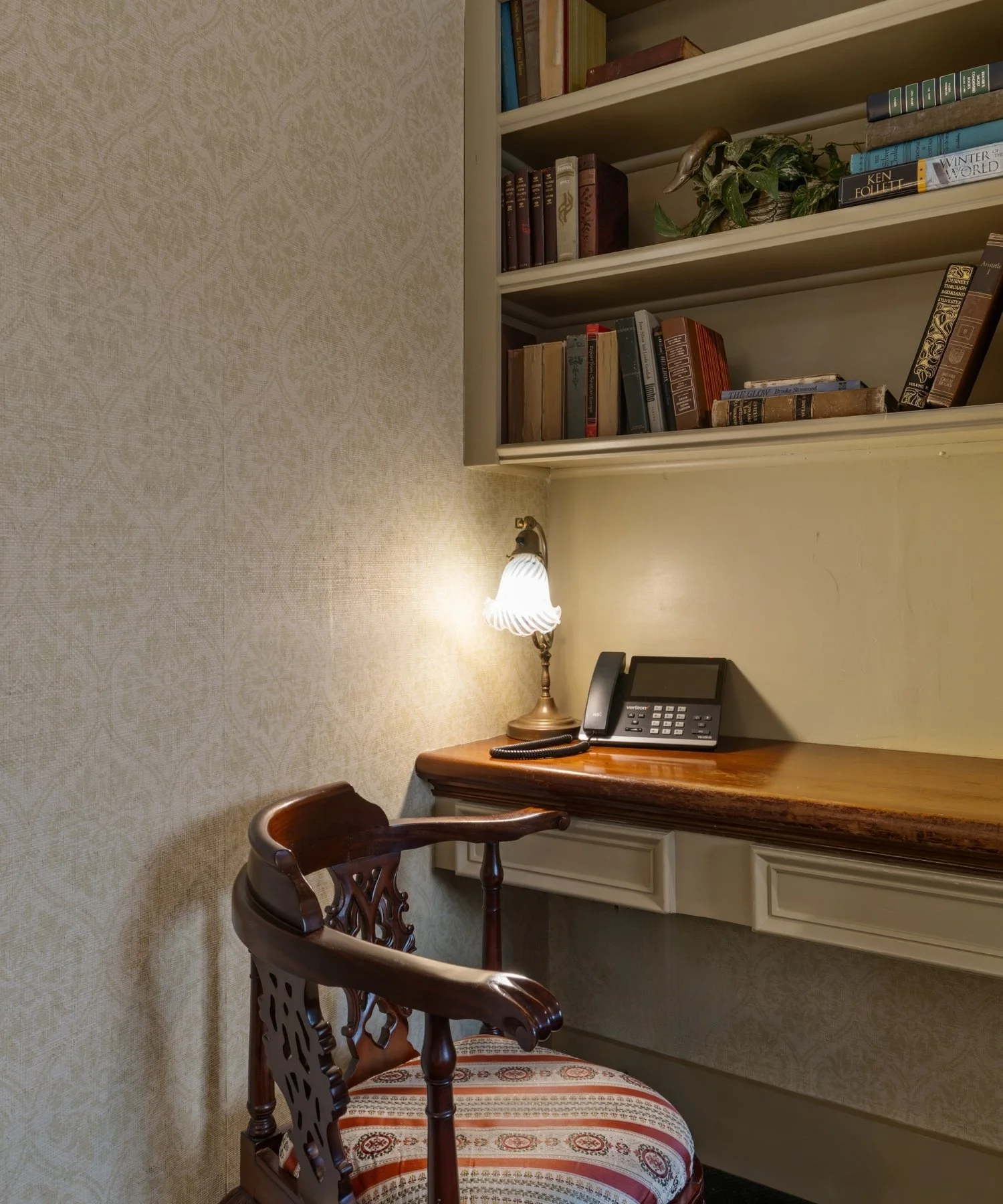 Wooden chair with carved backrest and upholstered seat in front of a desk with a landline phone, lamp, and a bookshelf with books and decorative items.