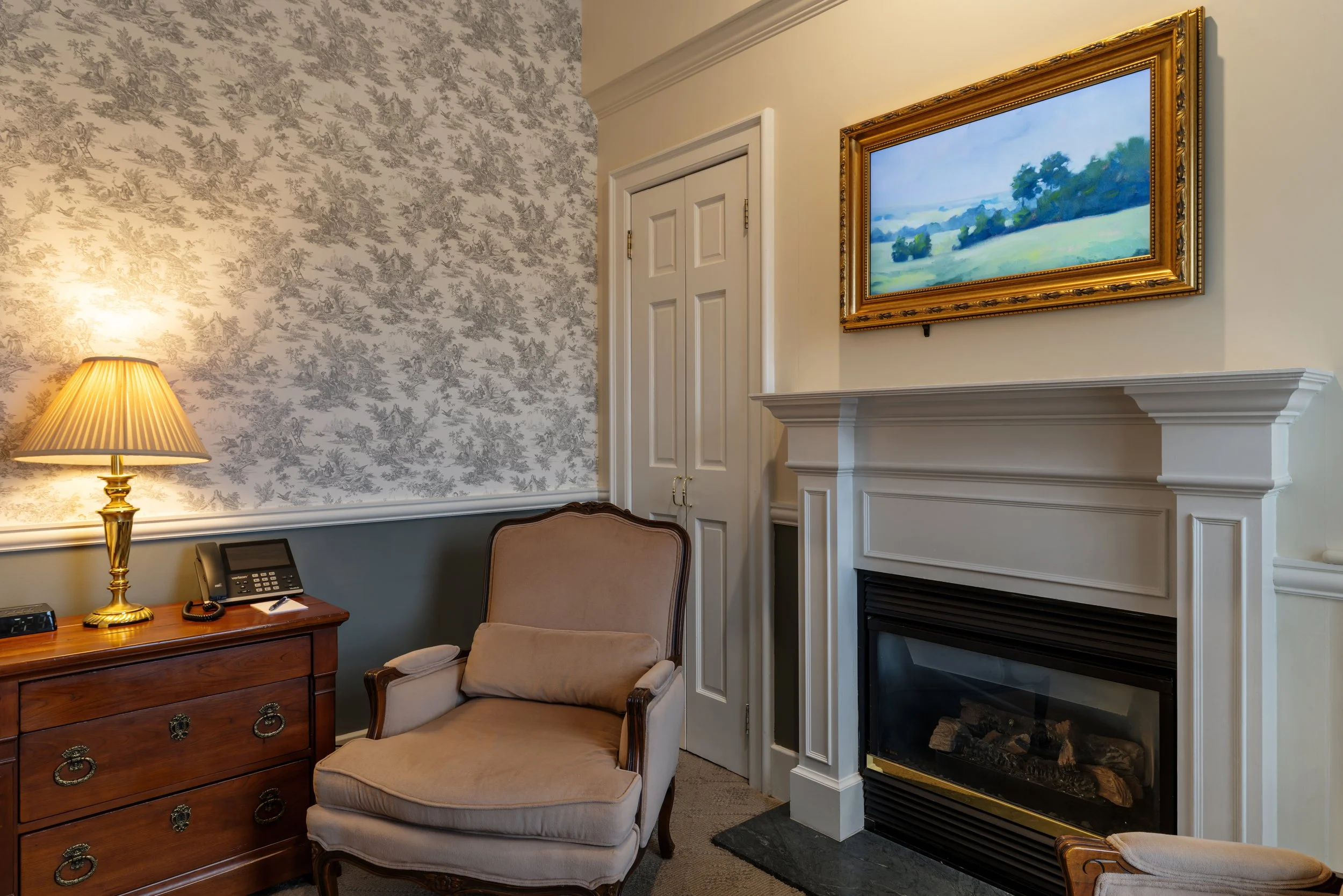 An interior room with a wooden desk, a lamp, a phone, and a chair. There is a fireplace with a decorative white mantle, a framed landscape painting, and floral wallpaper on one wall.