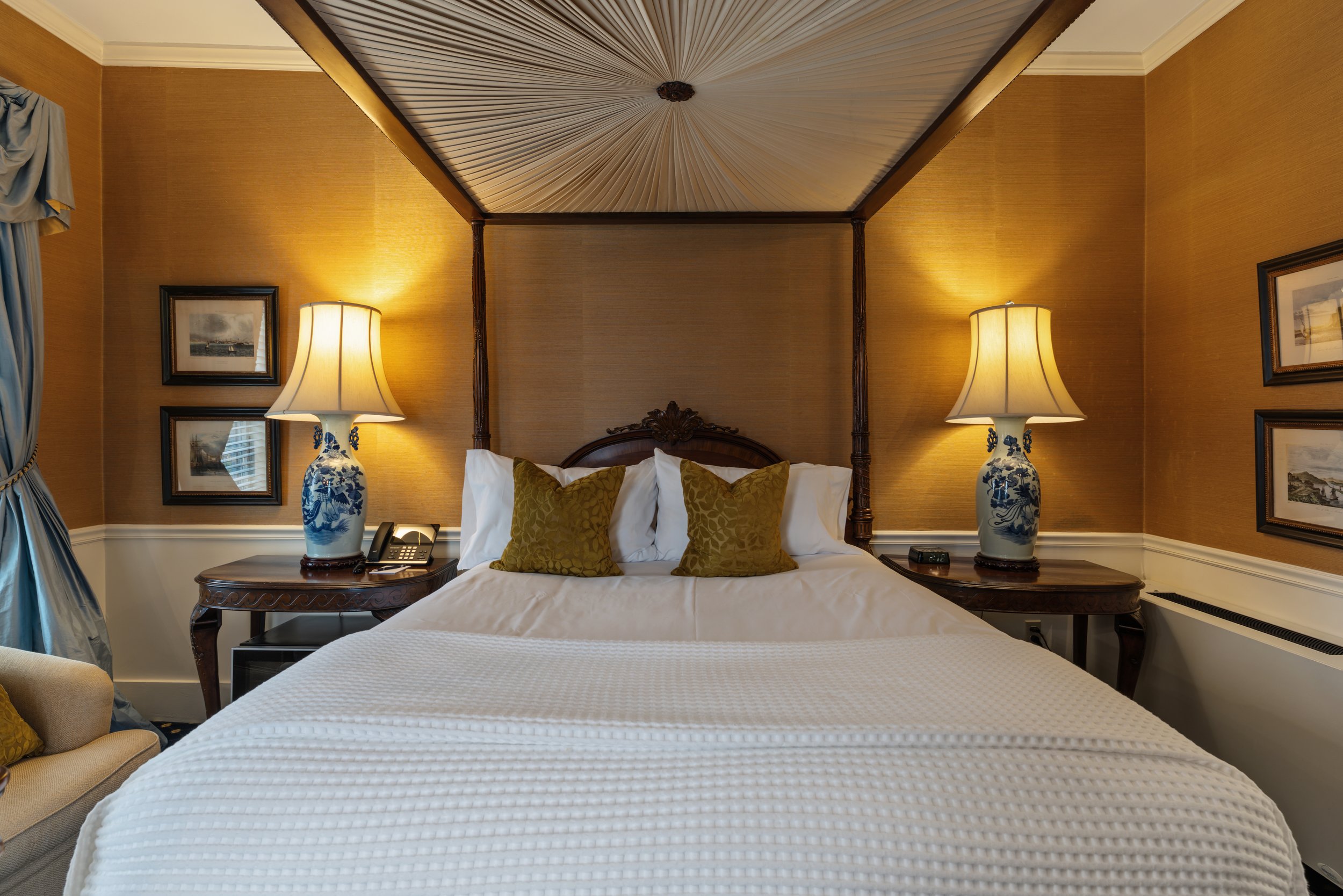 A hotel bedroom with a four-poster bed, bedside lamps, framed artwork, and a window with draped curtains.