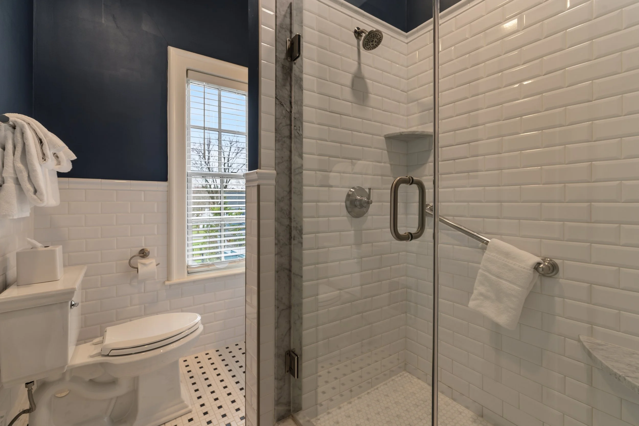 Bathroom with a white toilet, towel, window with blinds, and a walk-in shower with white subway tile walls and a glass door.
