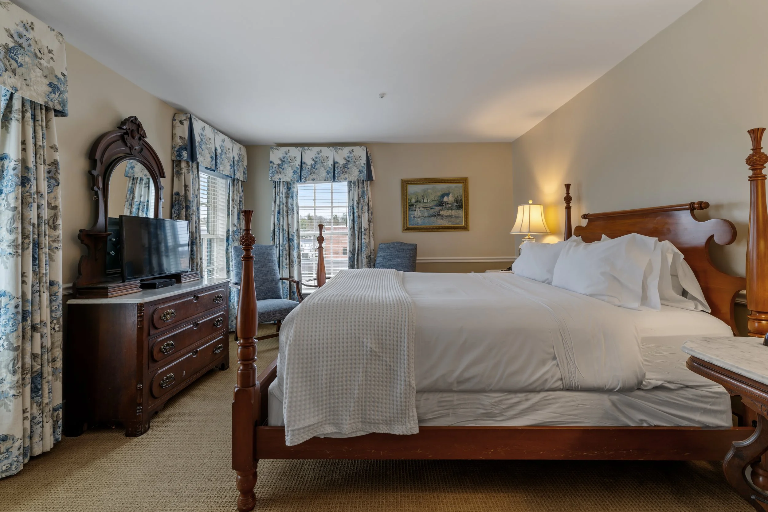 A hotel room with a wooden bed, white bedding, a lamp, two chairs, a framed painting, a flat-screen TV on a wooden dresser, and floral curtains.