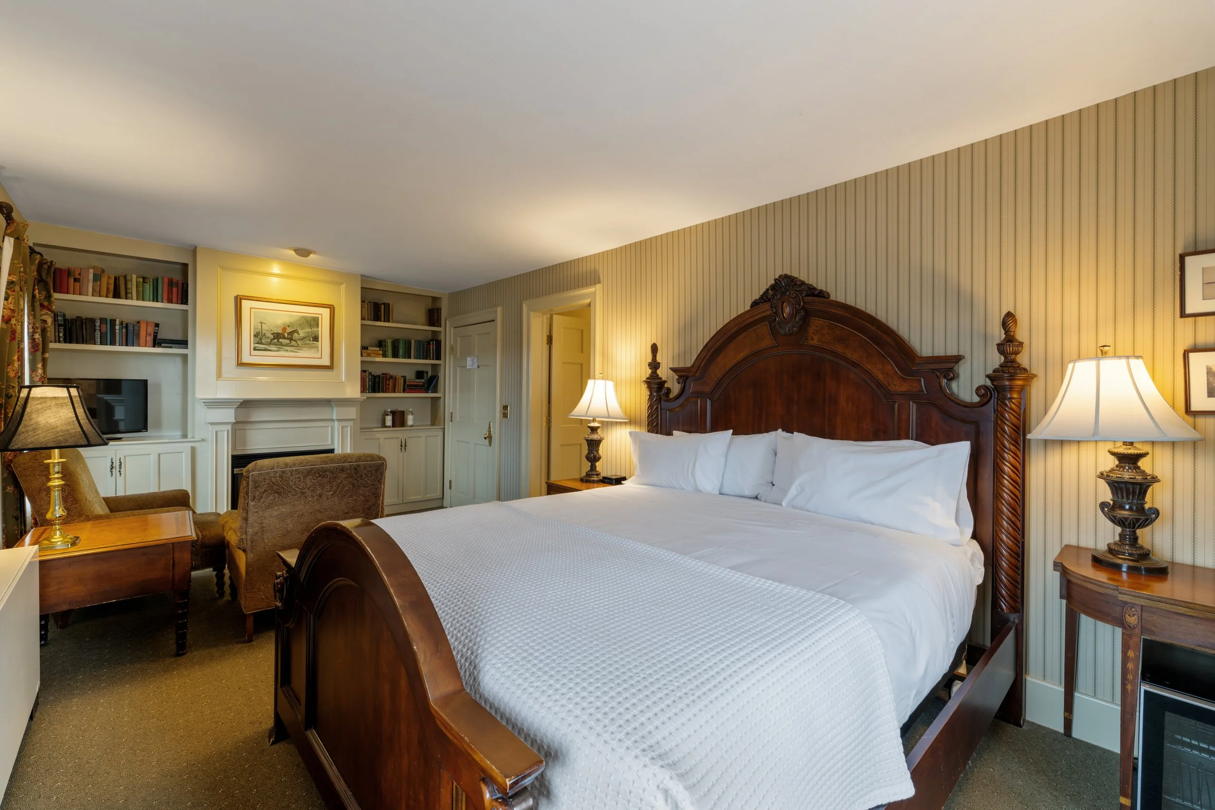 A hotel room with a wooden bed frame, white bedding, bedside lamps, and built-in bookshelves filled with books, artwork, and a small television.