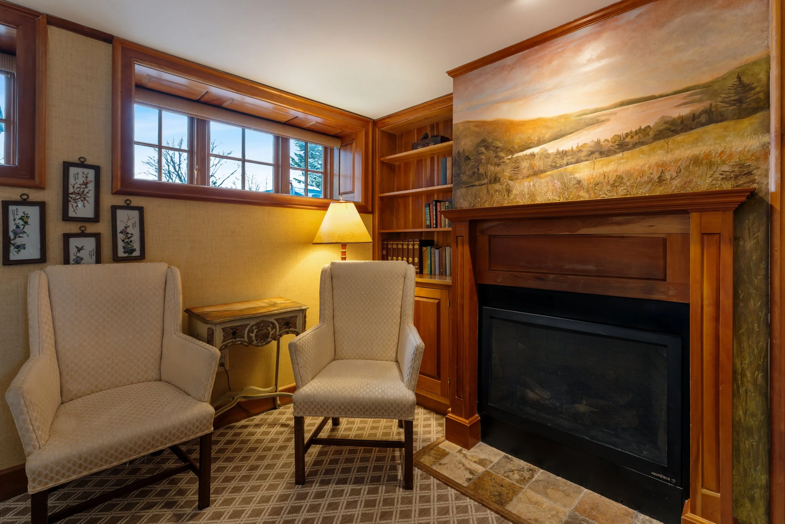 Living room area with beige armchairs, a small side table, a standing lamp, large windows, and a mural of a landscape above a fireplace.