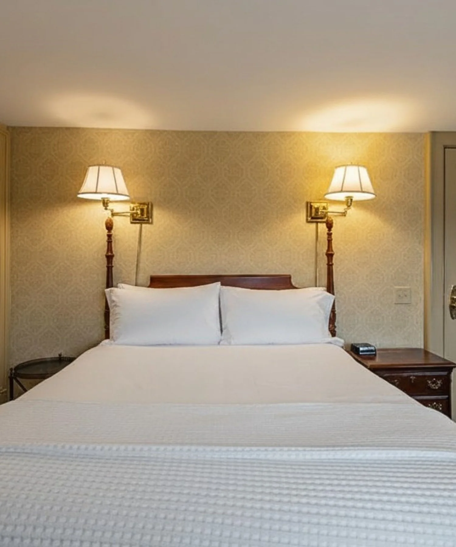 Hotel room with a bed, two wall-mounted lamps, a nightstand with a telephone, and patterned wallpaper.