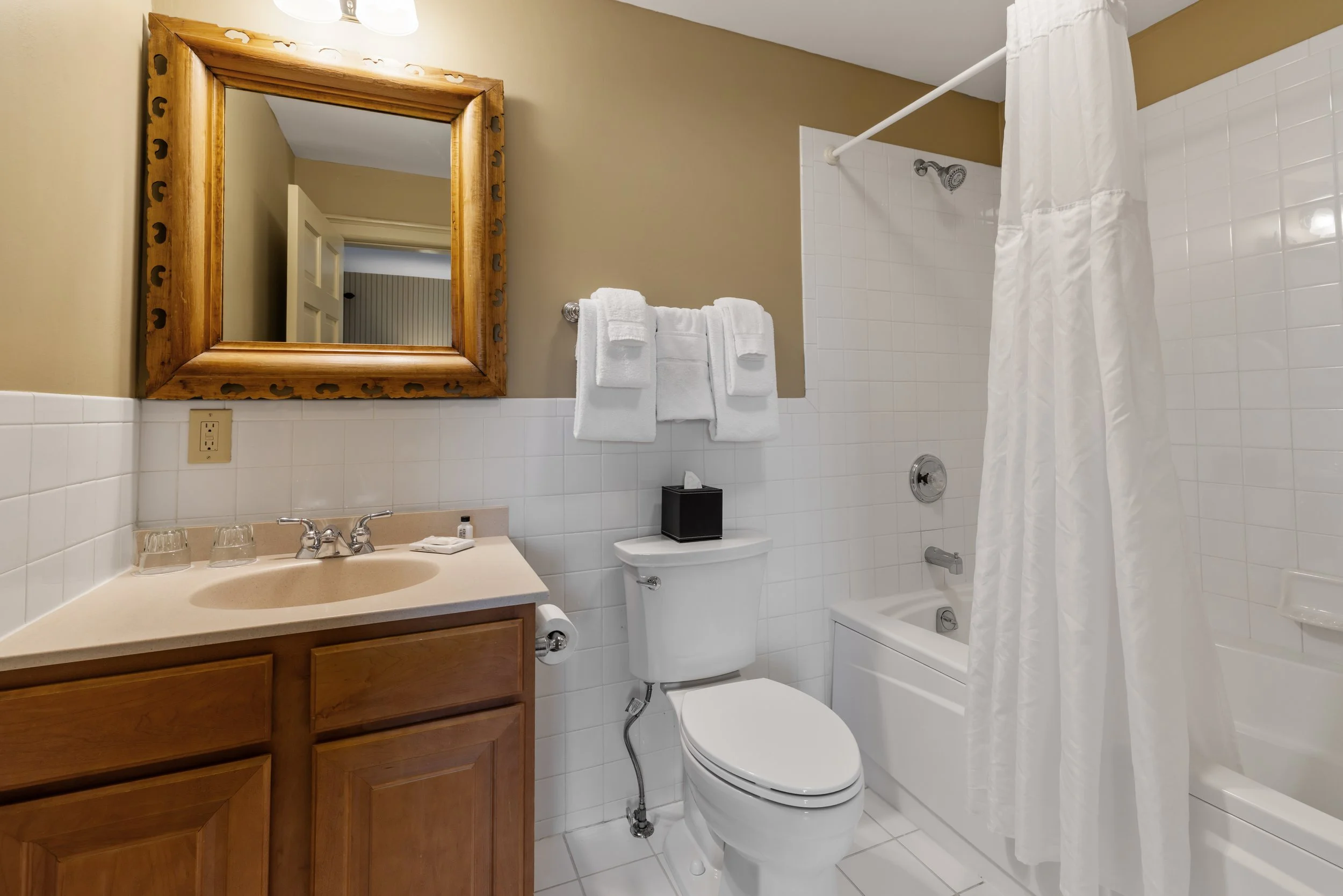 A small bathroom with a wooden framed mirror above the sink, a white towel rack with towels, a toilet, a bathtub with a shower curtain, beige walls, and white tiled walls.