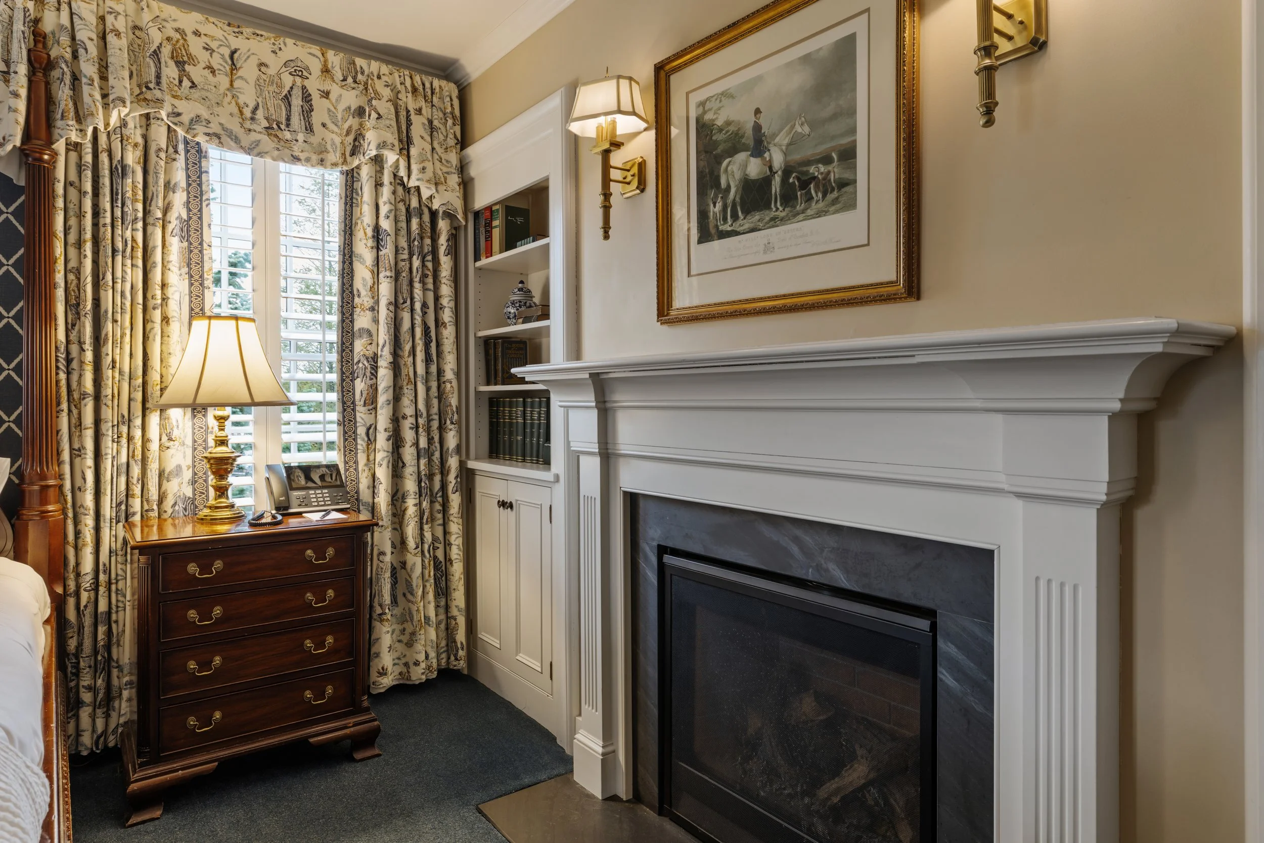 Interior of a room with a fireplace, framed artwork, bookshelf, a table with a lamp, and patterned curtains.