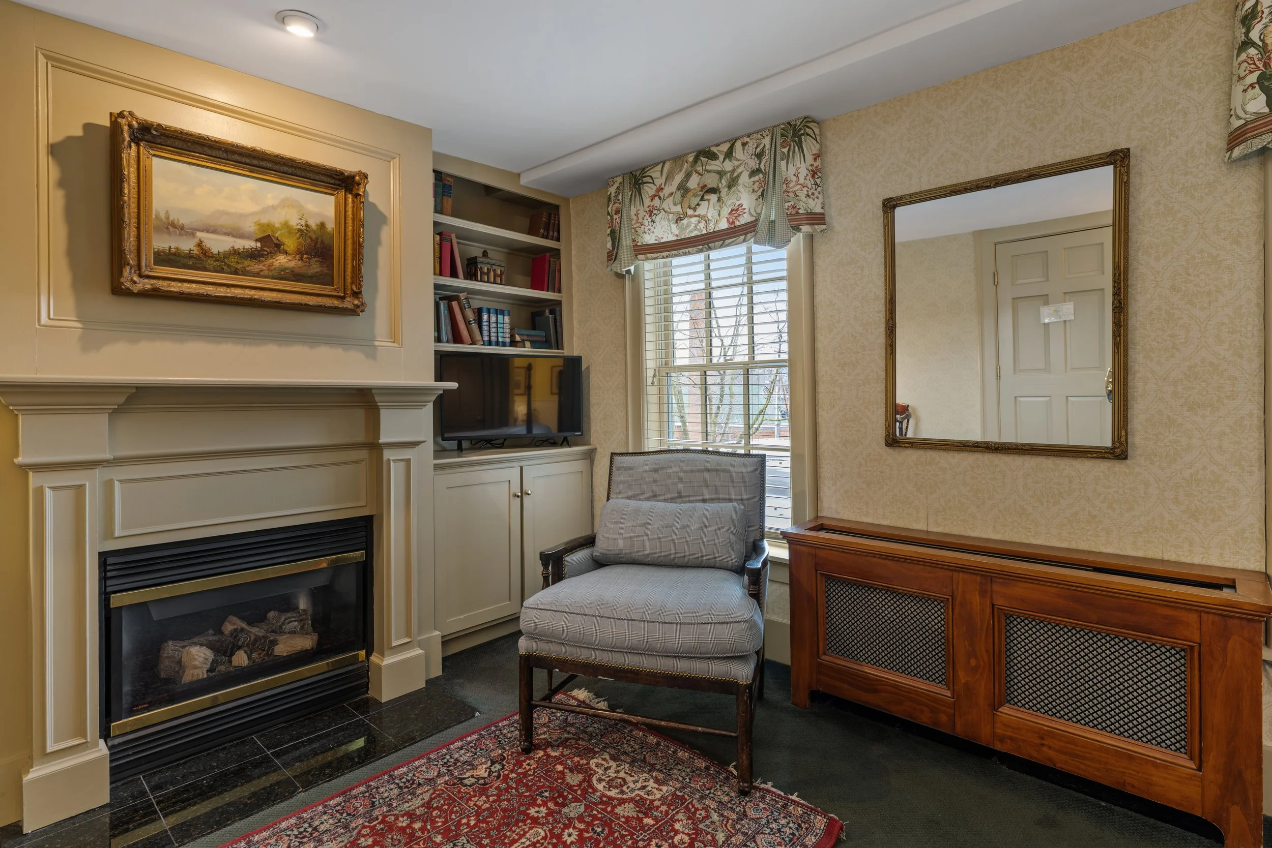 Living room corner with a fireplace, bookshelf, window with floral valance, mirror, armchair, wooden cabinet, and a rug.