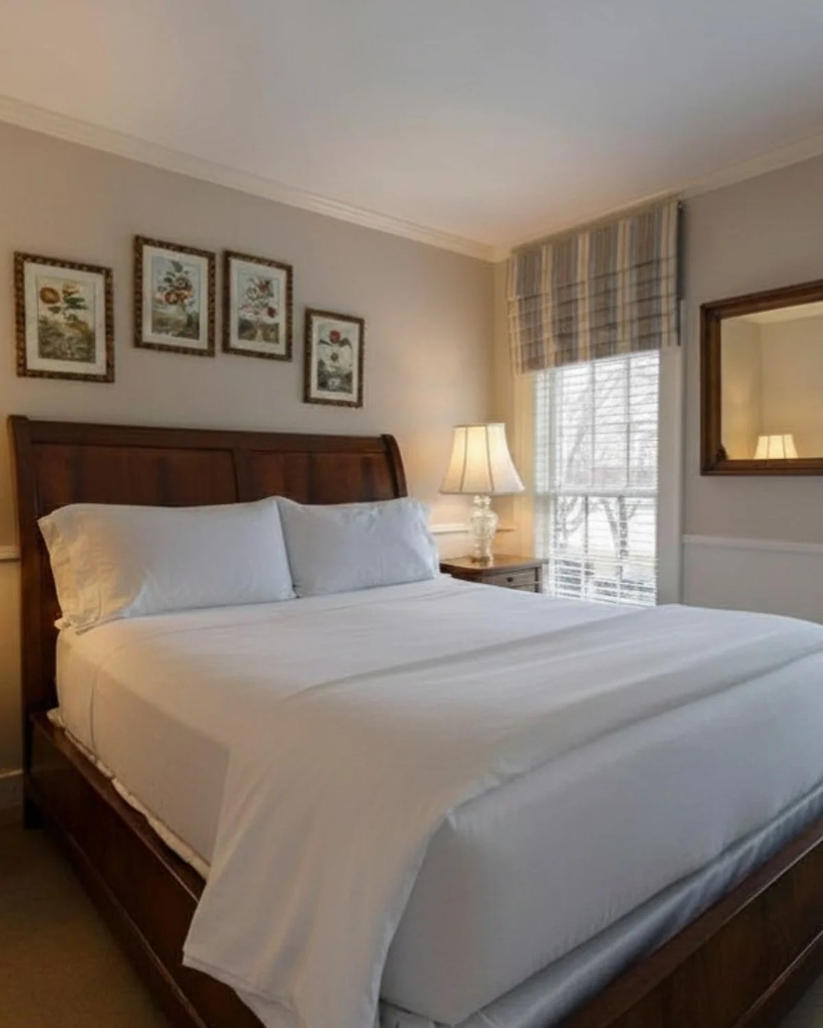 A cozy bedroom with a wooden bed frame, white bedding, a bedside table with a lamp, framed artwork on the wall, and a window with blinds and a plaid valance.