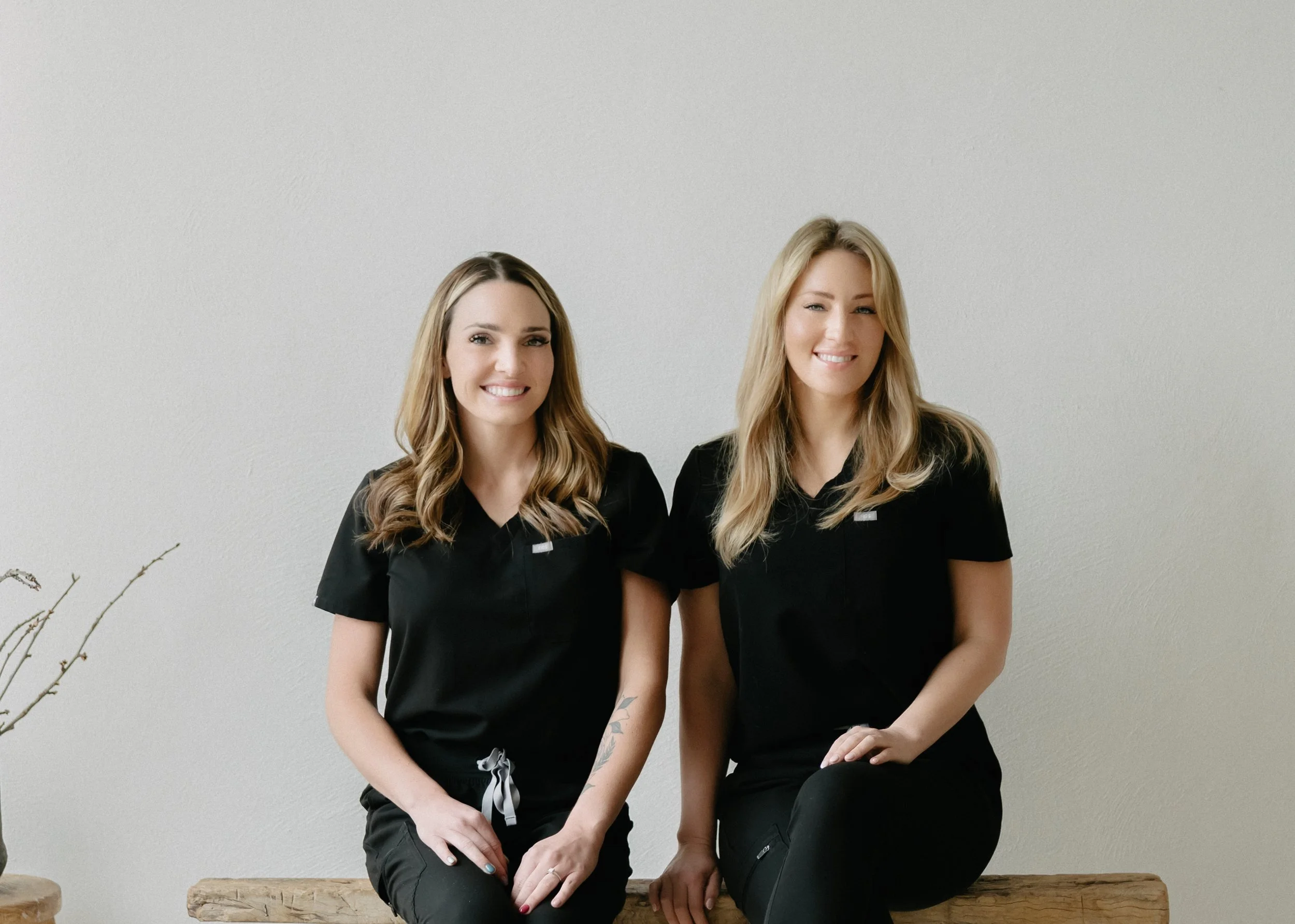 Two smiling women with blonde hair and black scrubs sitting on a wooden bench in front of a plain white wall.