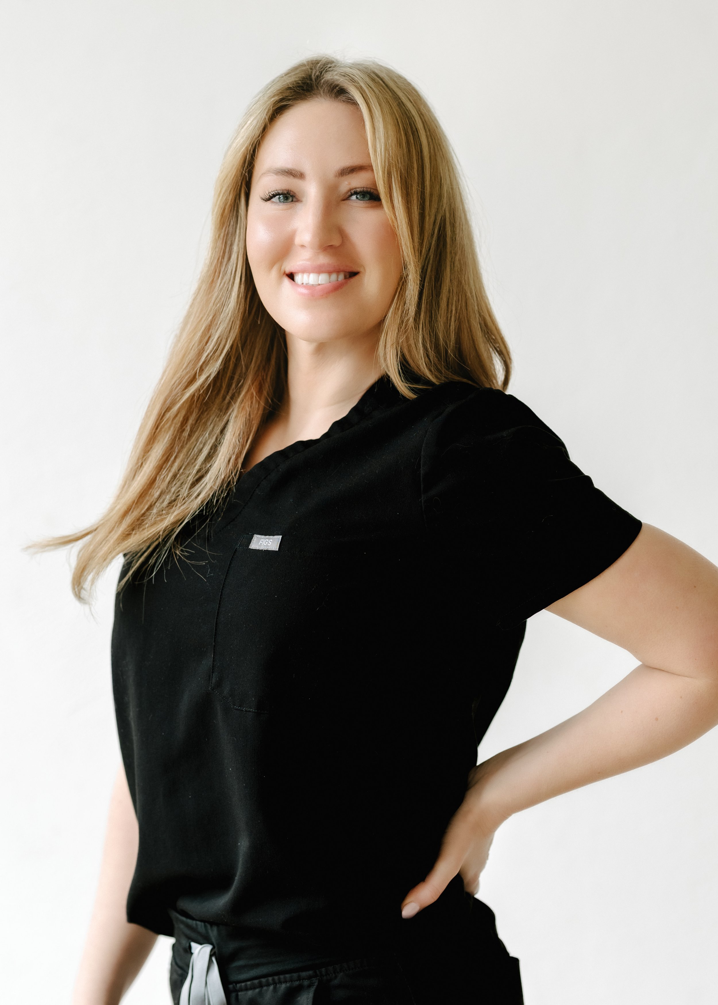 Young woman with long blonde hair smiling and wearing a black medical uniform, standing against a plain white background.
