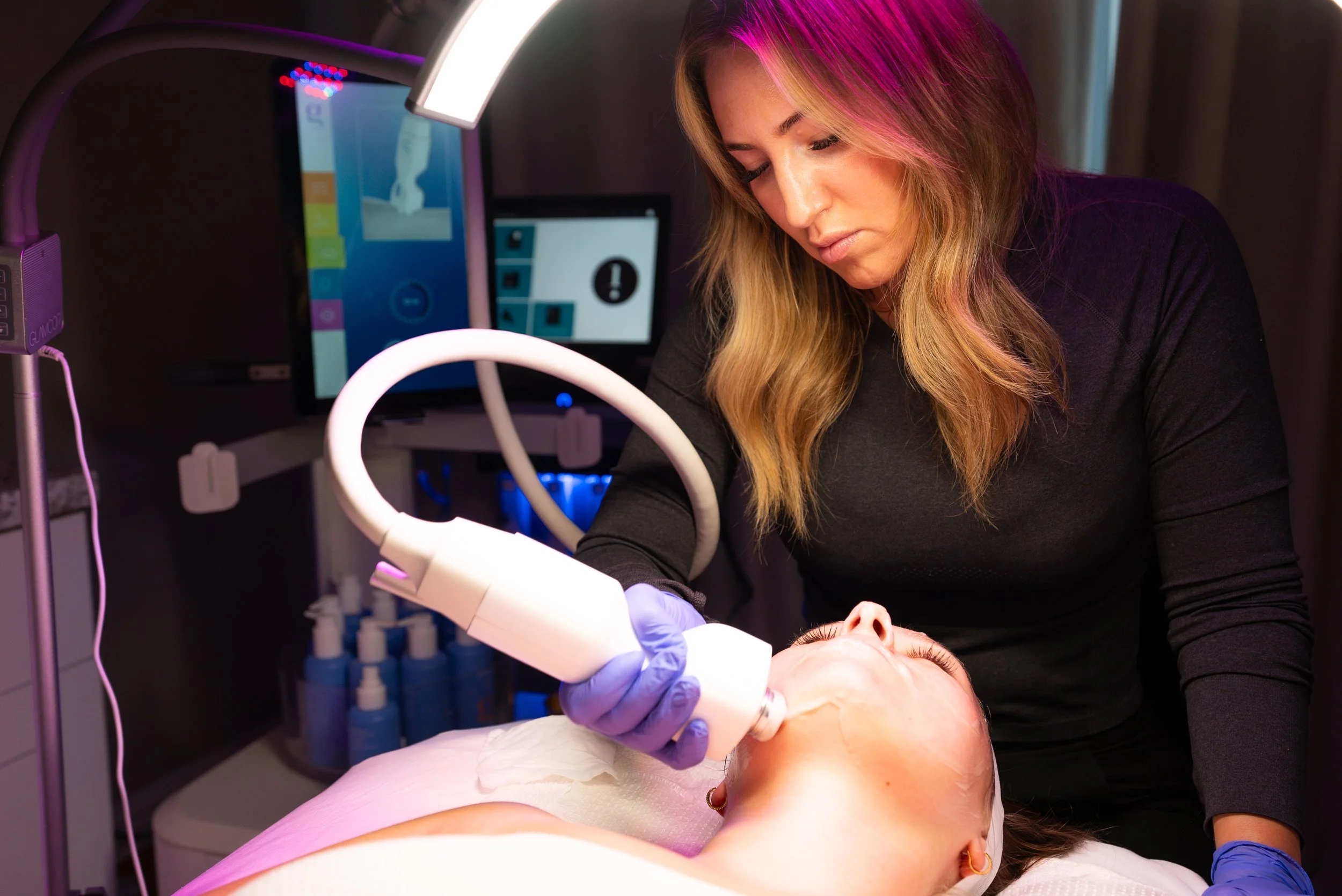 A woman performing a facial treatment using a medical device on a woman's face in a clinical setting.