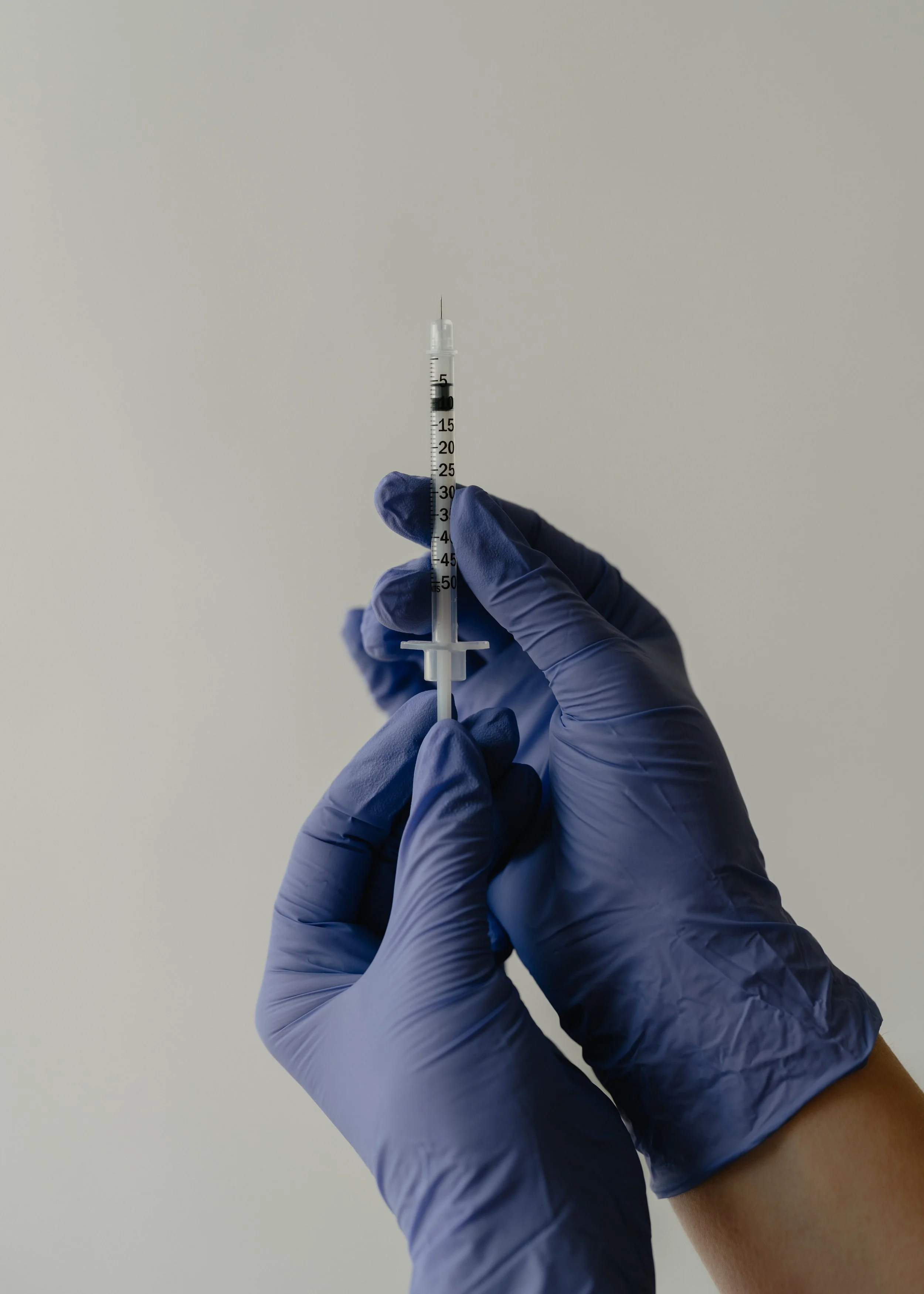 Hands wearing blue gloves holding a syringe filled with a clear liquid against a plain background.