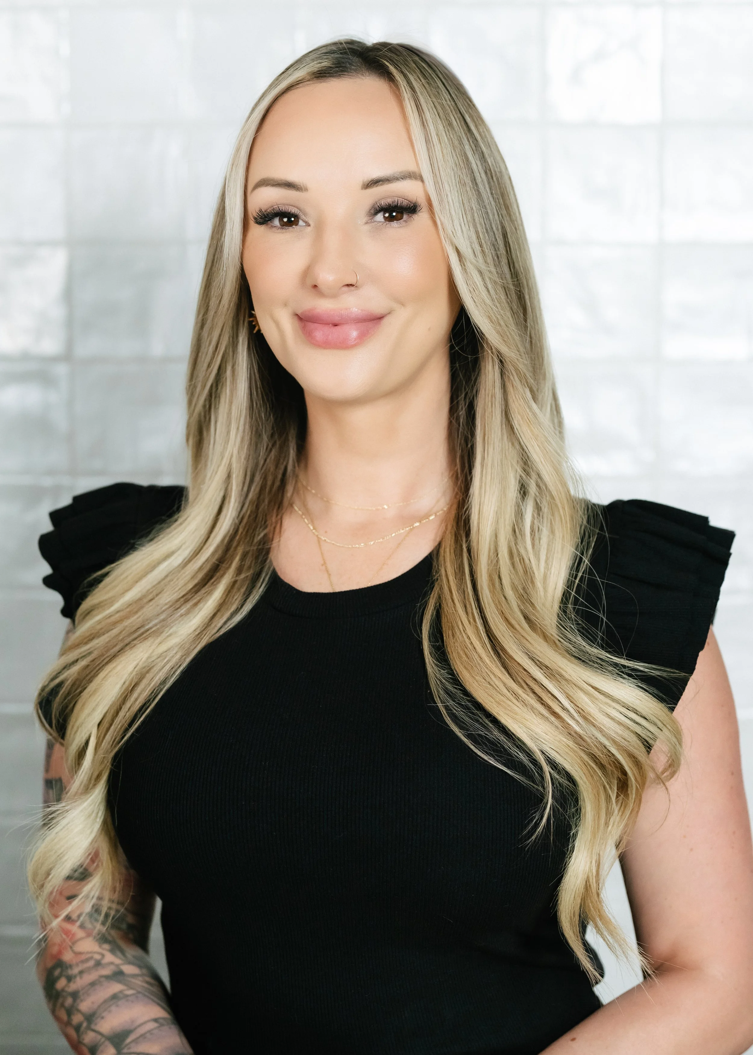A woman with long wavy blonde hair wearing a black top with ruffled sleeves, gold jewelry, and a nose piercing, smiling in front of a white tiled wall.