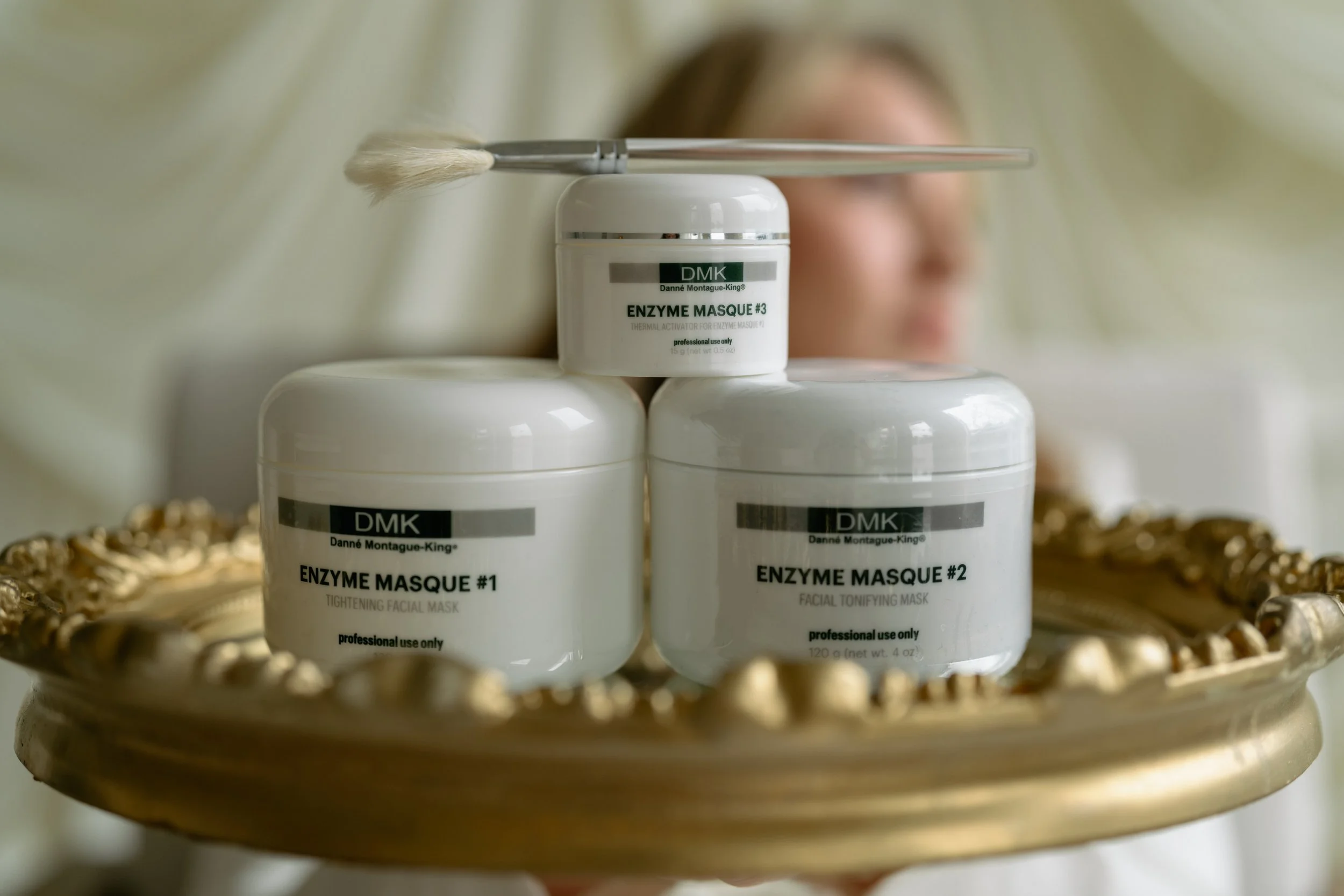 Three containers of enzyme masks placed on a decorative tray with a blurred face of a person in the background.