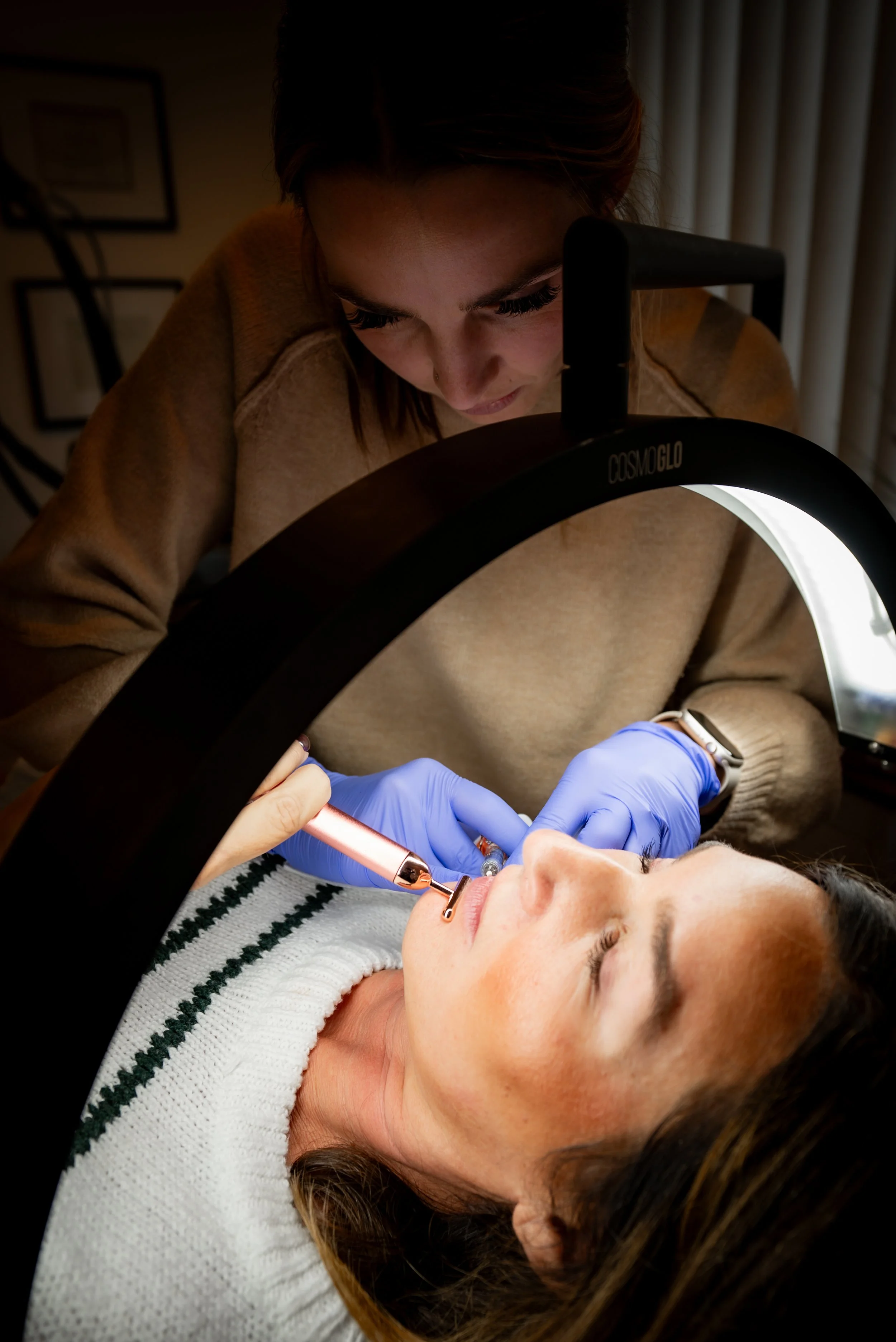 A woman is receiving a cosmetic procedure on her lips while lying down, with a technician using a device and wearing blue gloves, under a specialized light