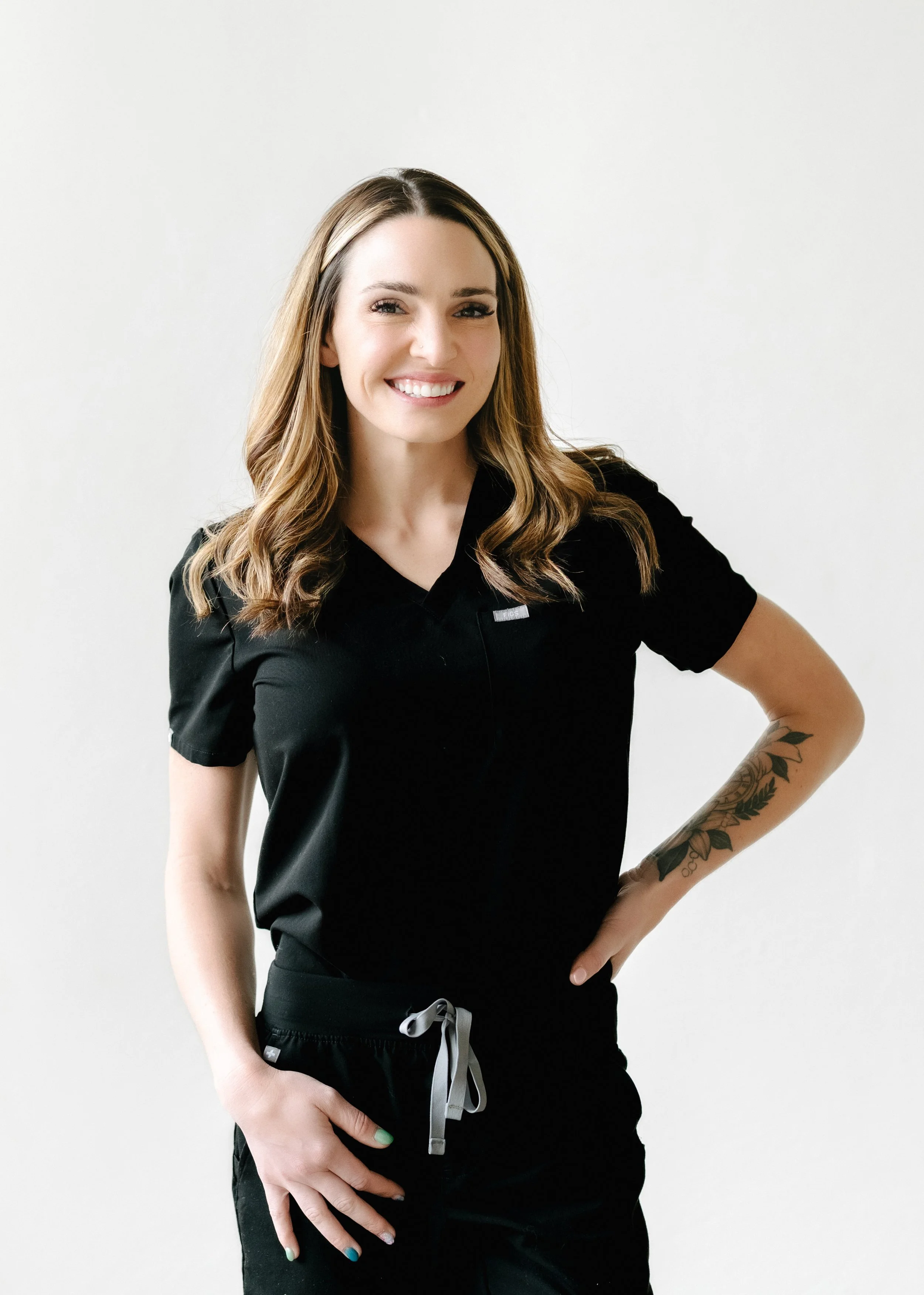 A smiling woman with wavy brown hair, wearing a black uniform with a small name tag, standing against a plain white background.