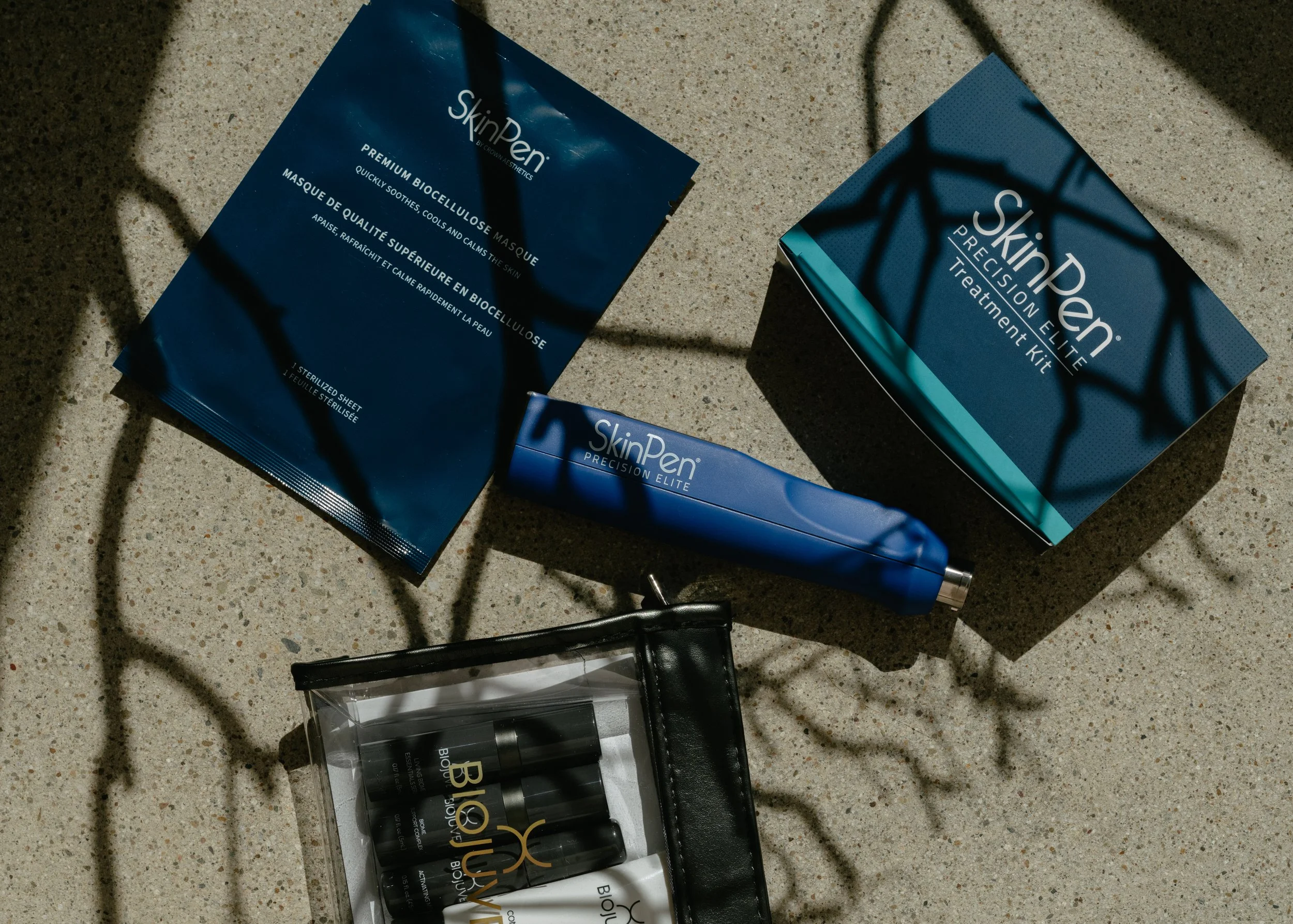 Skinceuticals skincare products including SkinPen Precision Elite treatment kit, a single use mask packet, and a small pouch with several vials, all displayed on a textured surface with shadows of leafless branches.