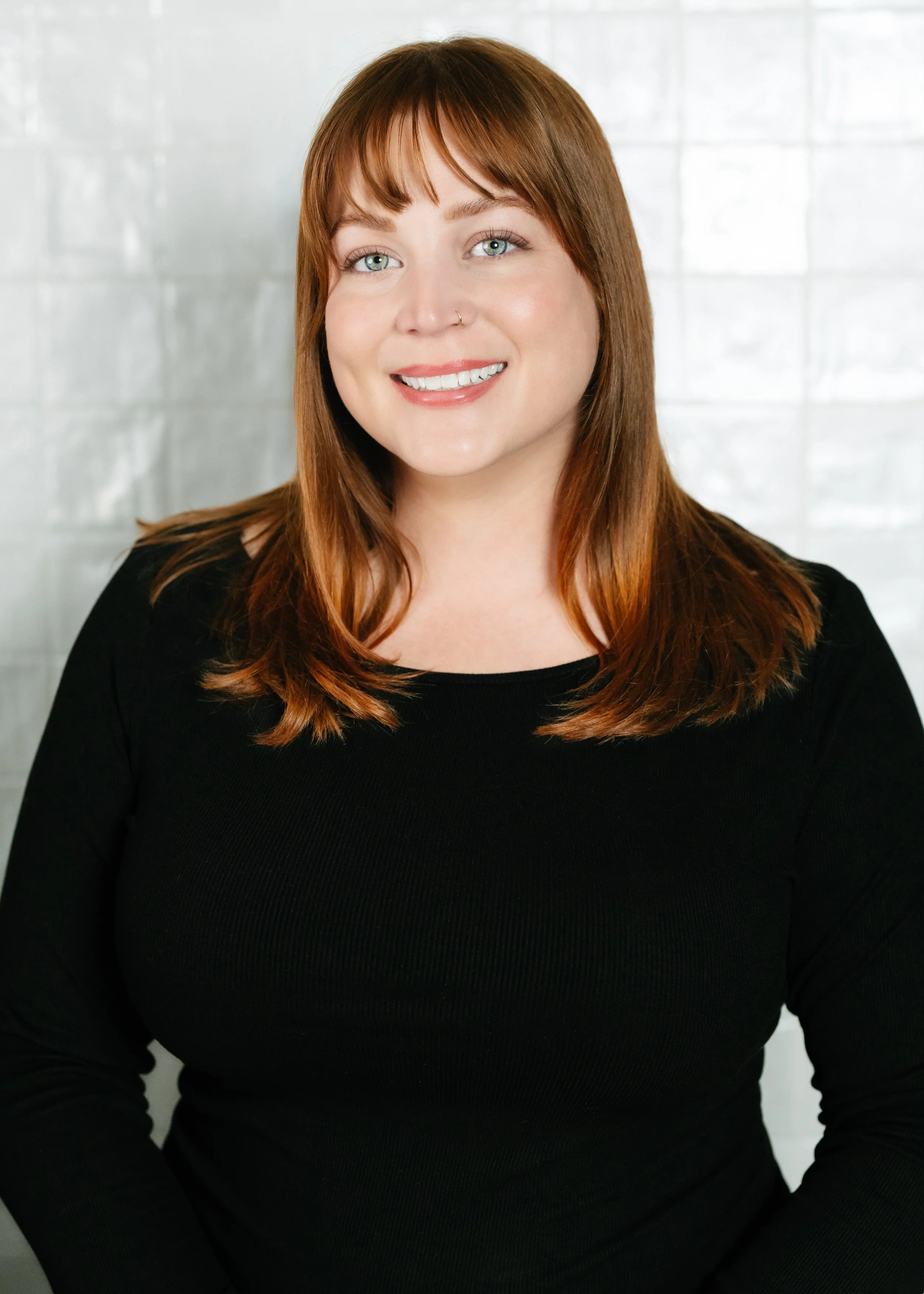 A woman with red hair, blue eyes, and a nose ring, smiling while wearing a black top, standing in front of a light-colored brick wall.