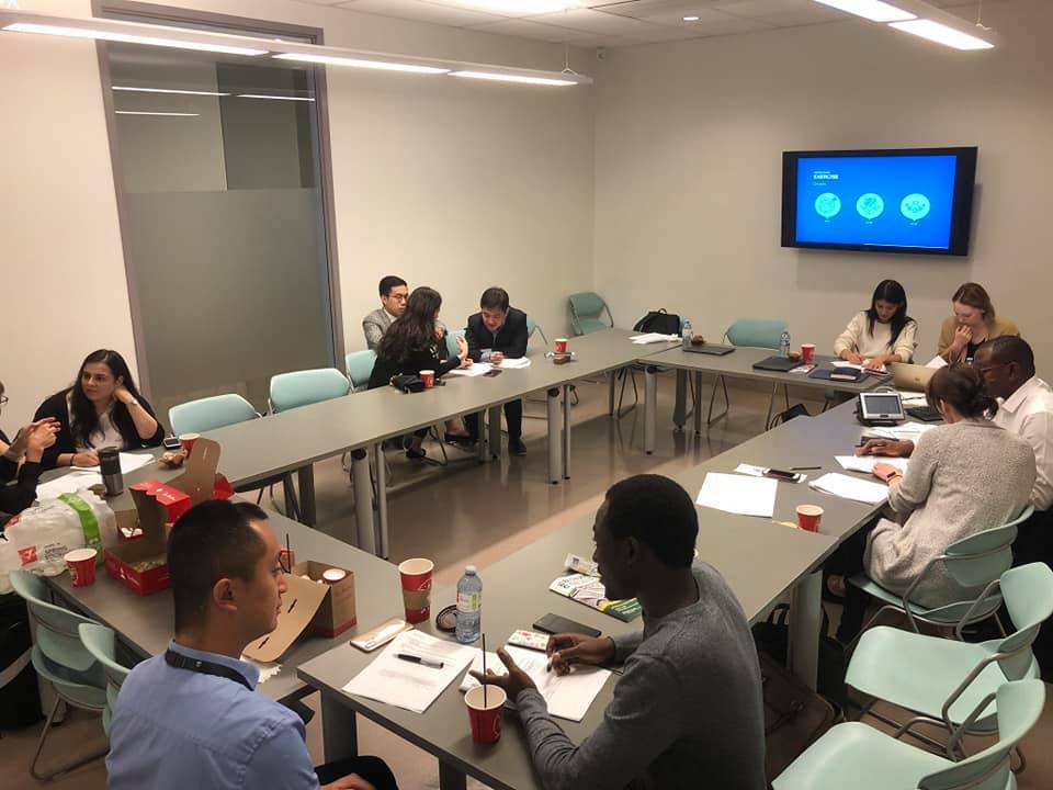 Business meeting in a conference room with people sitting around a U-shaped table, some with laptops, notebooks, and cups of coffee, while a large screen displays a presentation.