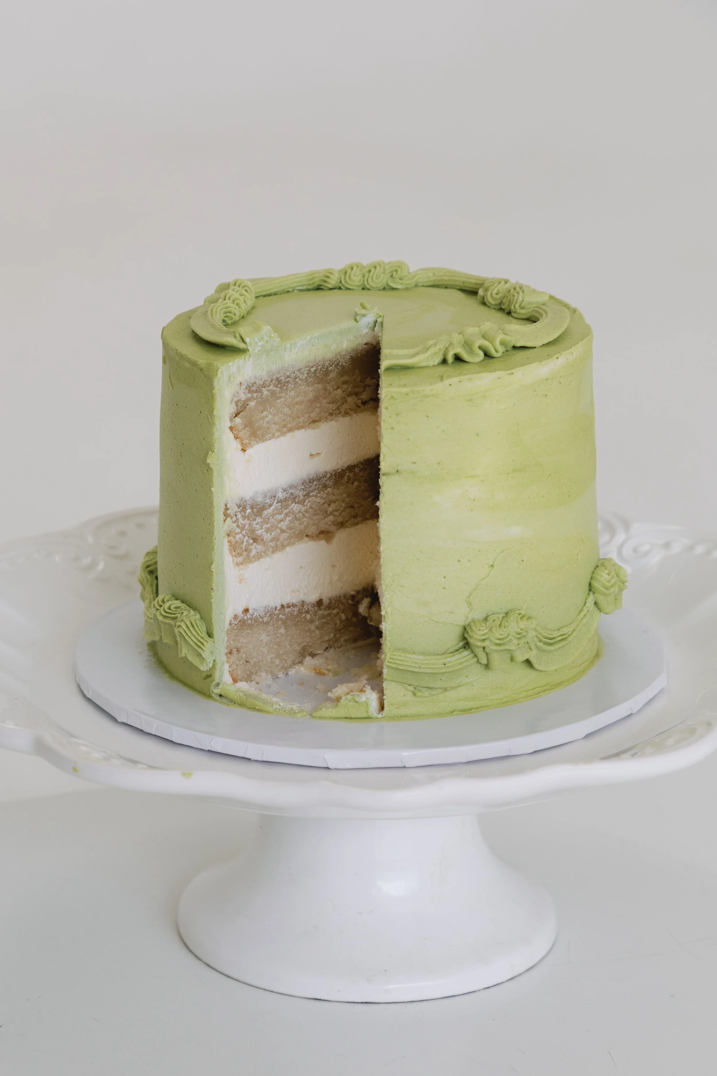 A partially sliced, three-layer vanilla cake with green frosting and piped decorations on top, presented on a white cake stand.