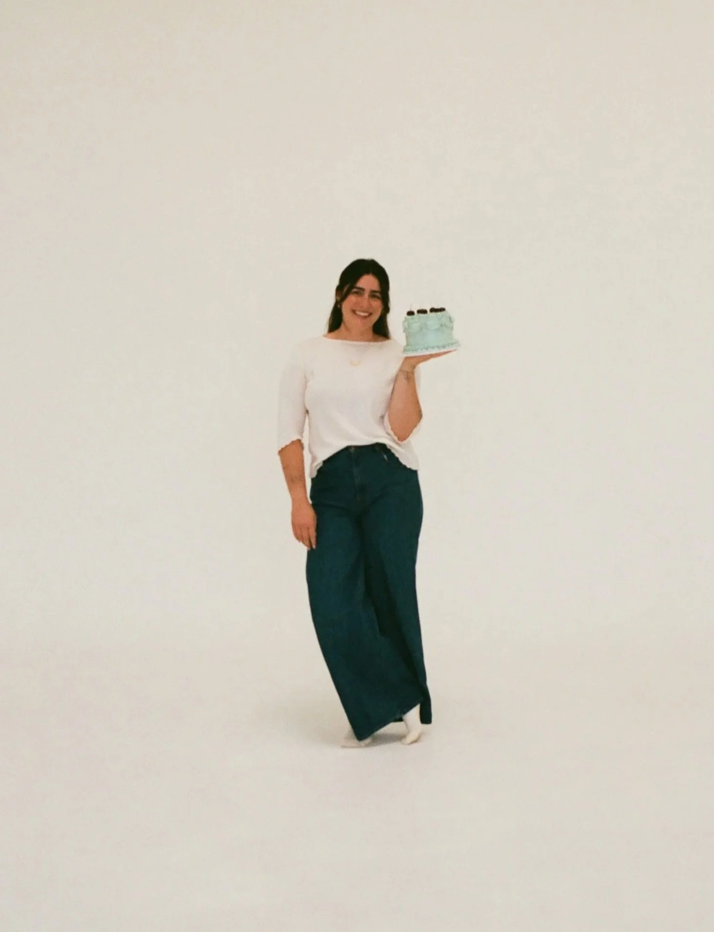 A photo of Grayce with long dark hair wearing a white top and dark wide-leg pants, smiling while holding a light blue birthday cake with dark cherries on top against a plain white background.