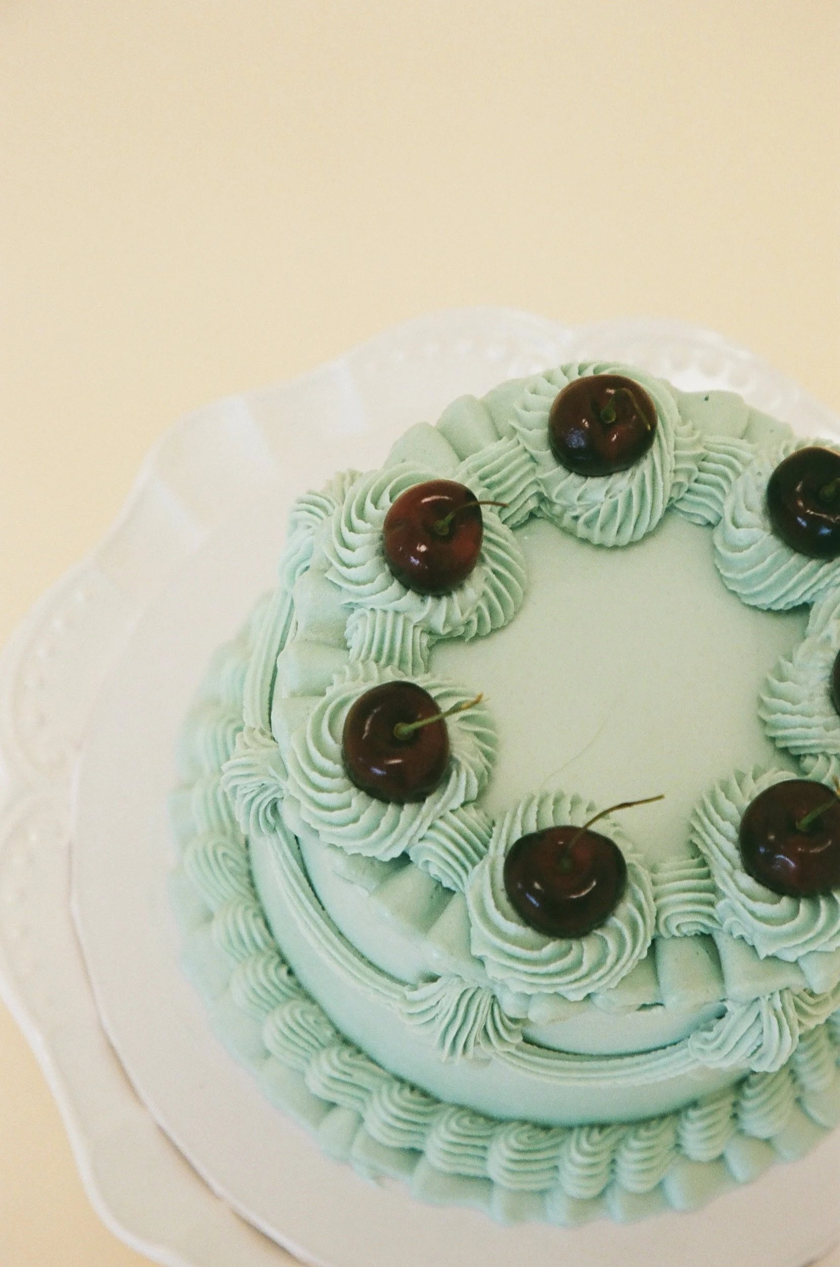 A photo of a round, light blue frosted birthday cake, topped with cherries and decorative piped frosting borders.