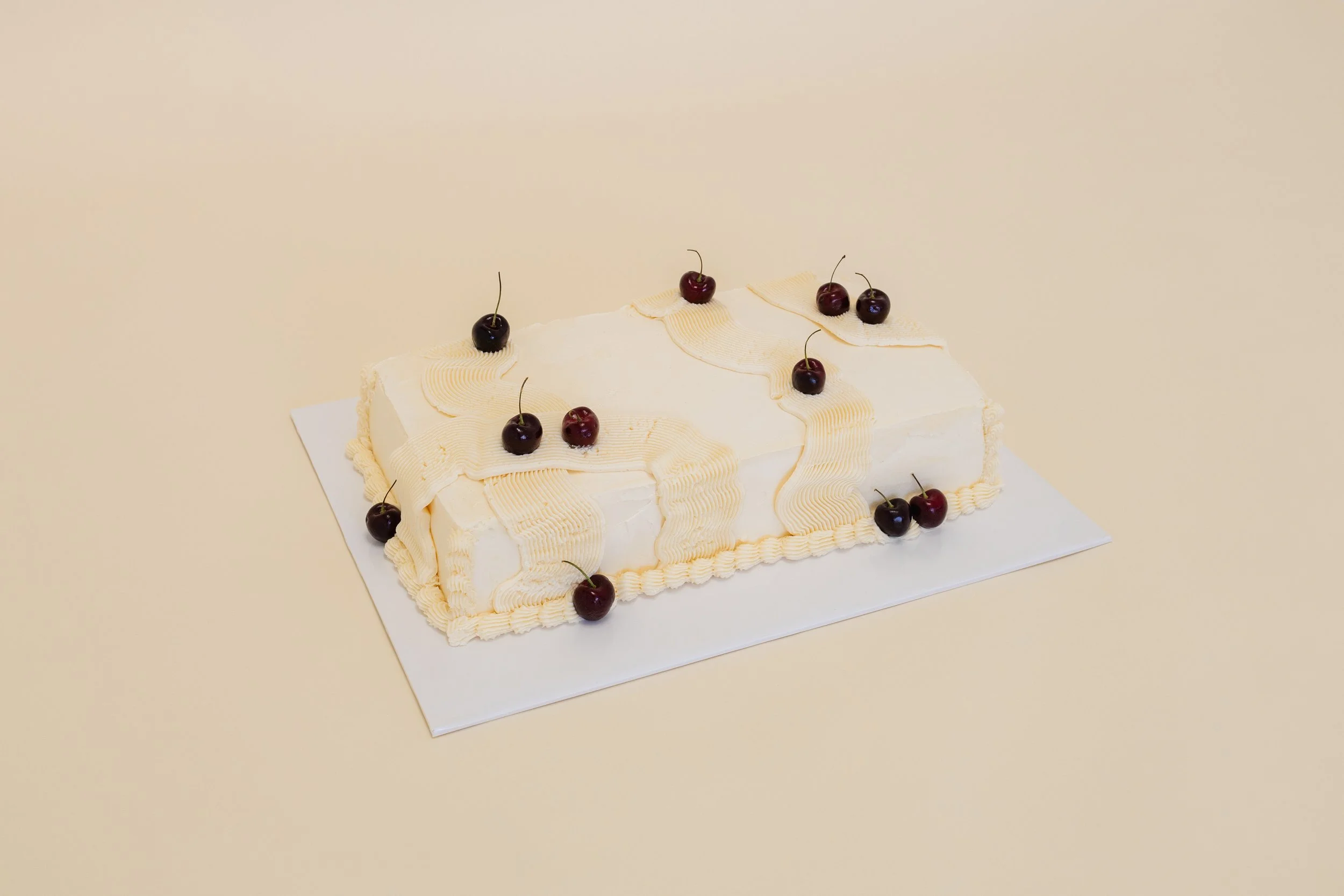 A rectangular cake with white frosting and yellow piping design, decorated with cherries on top, set on a white surface against a beige background.