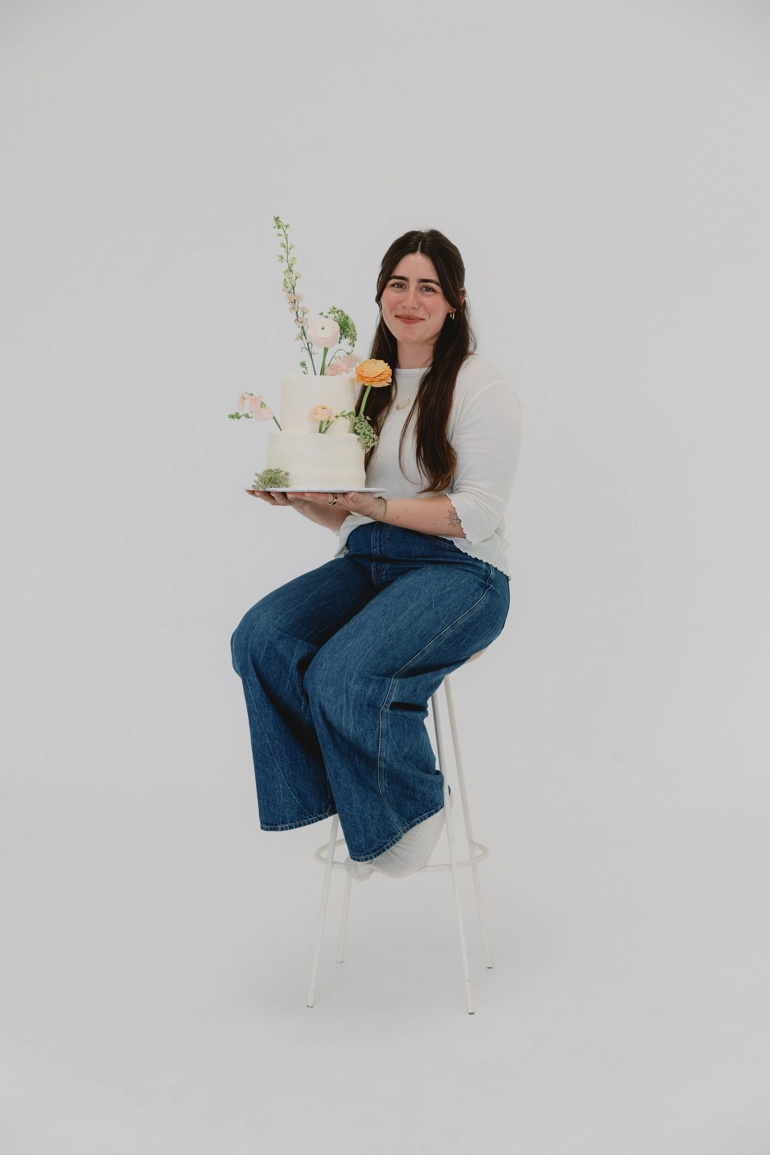 A photo of Grayce, with long brown hair, wearing a white long sleeve shirt and blue jeans, sitting on a high stool, holding a white cake decorated with real flowers, against a plain white background.