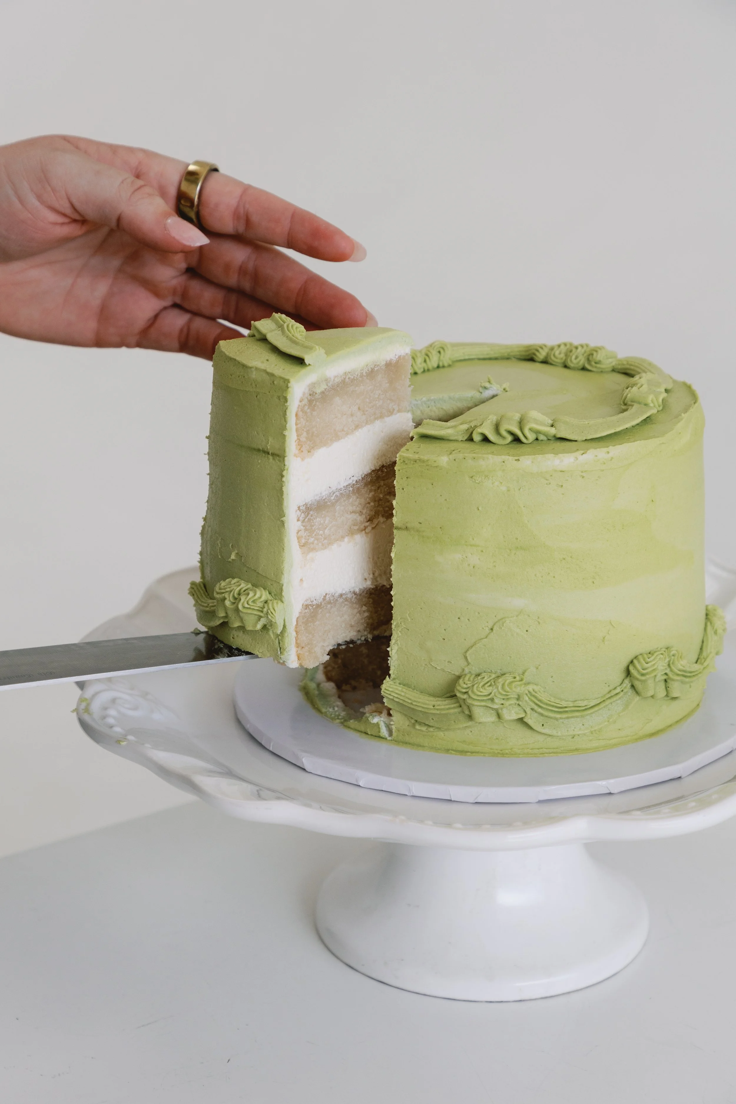 A person is cutting a slice from a green frosted layered cake on a white cake stand.
