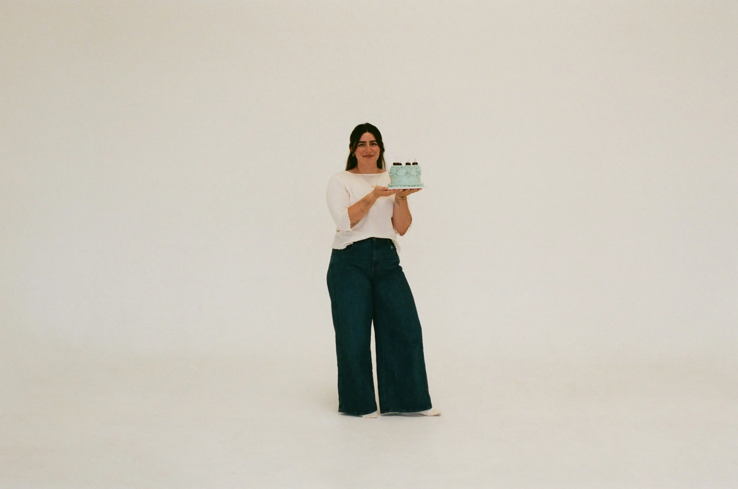 A photo of Grayce in white top and wide-leg jeans holding a birthday cake with blue frosting and cherries on top, standing in a plain white room.