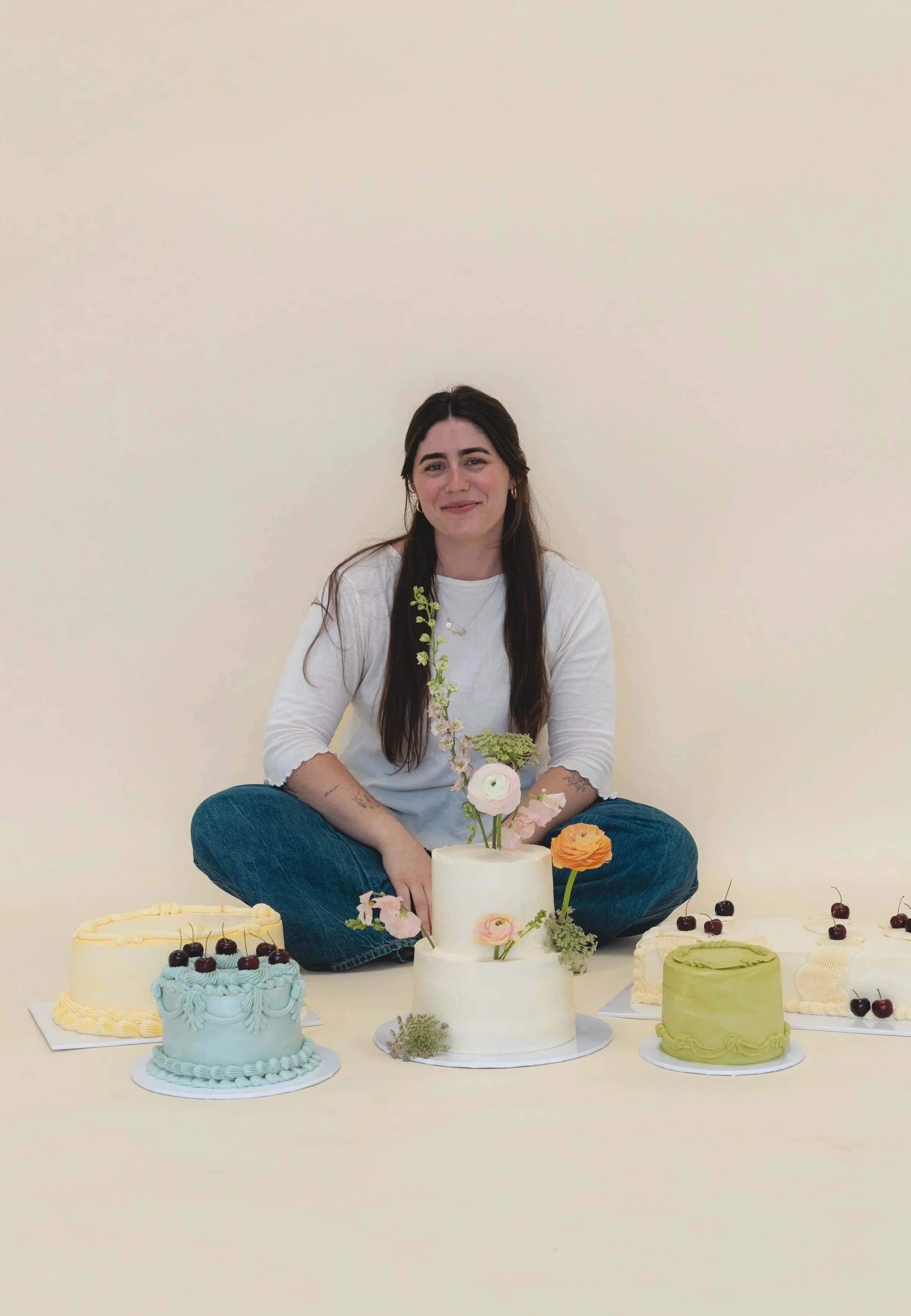 Grayce sitting cross-legged behind a variety of decorated cakes with flowers and cherries on a cream-colored background.