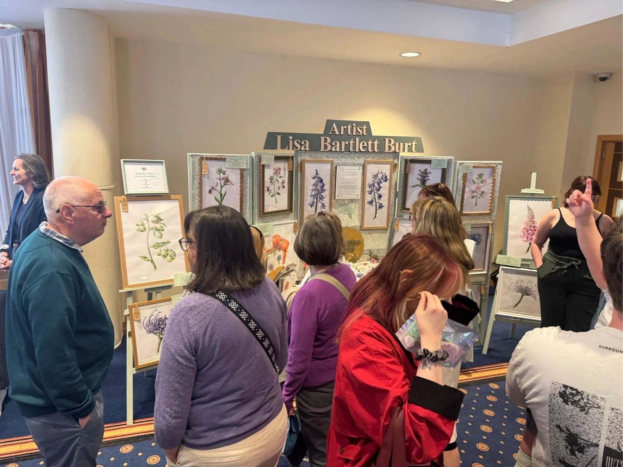 People attending an art exhibit display, titled "Lisa Bartlett Burt," featuring floral botanical illustrations and framed artwork, in a gallery with beige walls and blue carpeted floor.