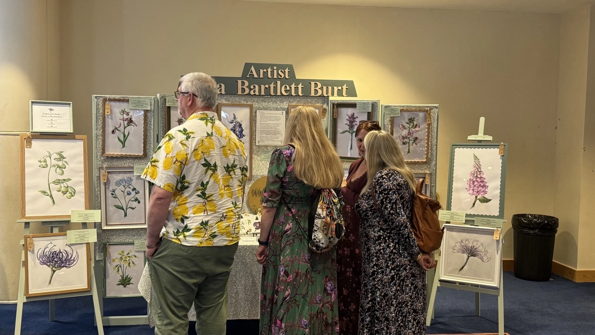 People viewing framed pressed flower art pieces at an art display titled 'Artist La Bartlett Burt'.