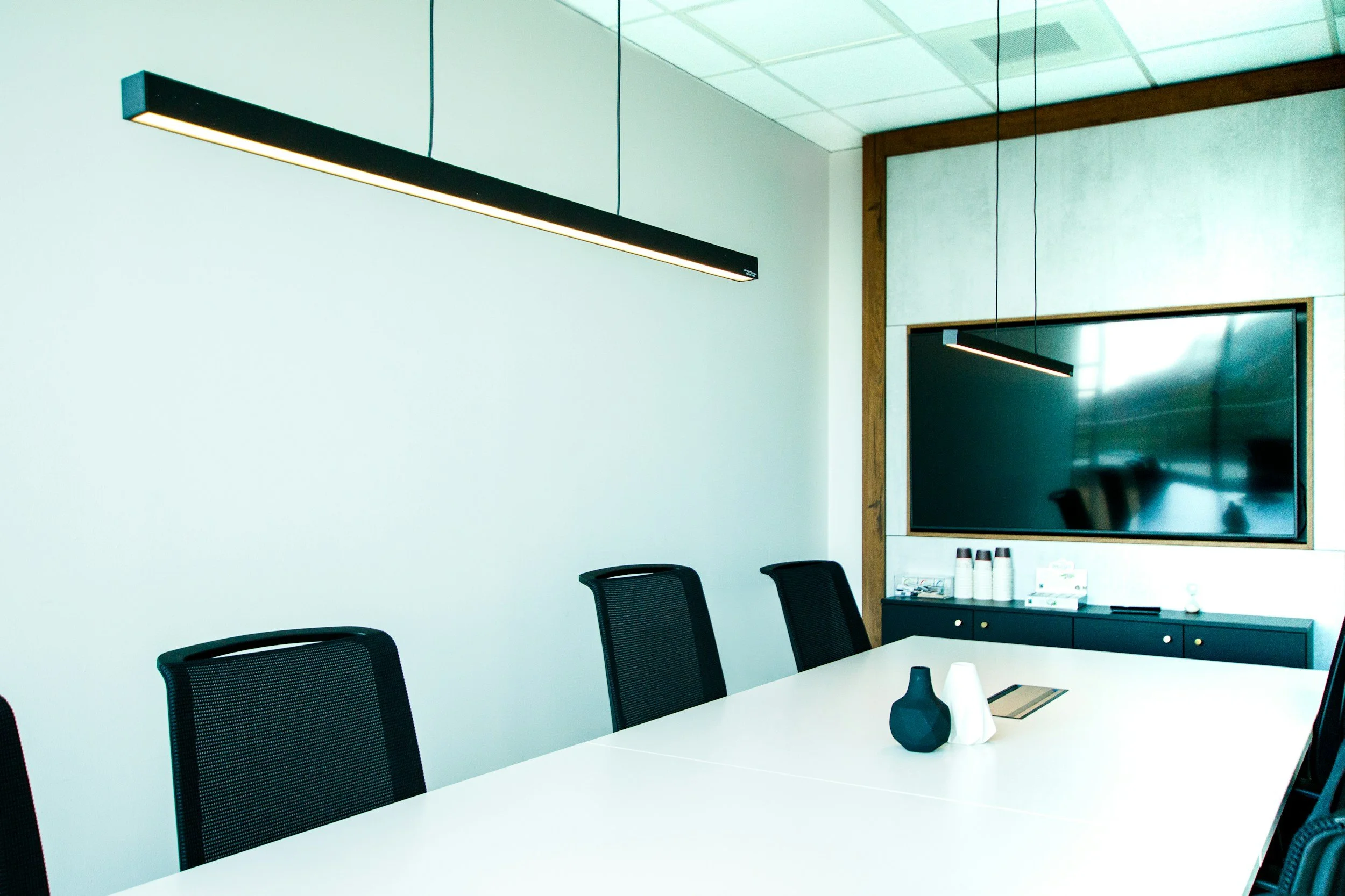 Empty modern conference room with white table, black chairs, black pendant lights, a large flat-screen TV, and a black cabinet with white bottles.