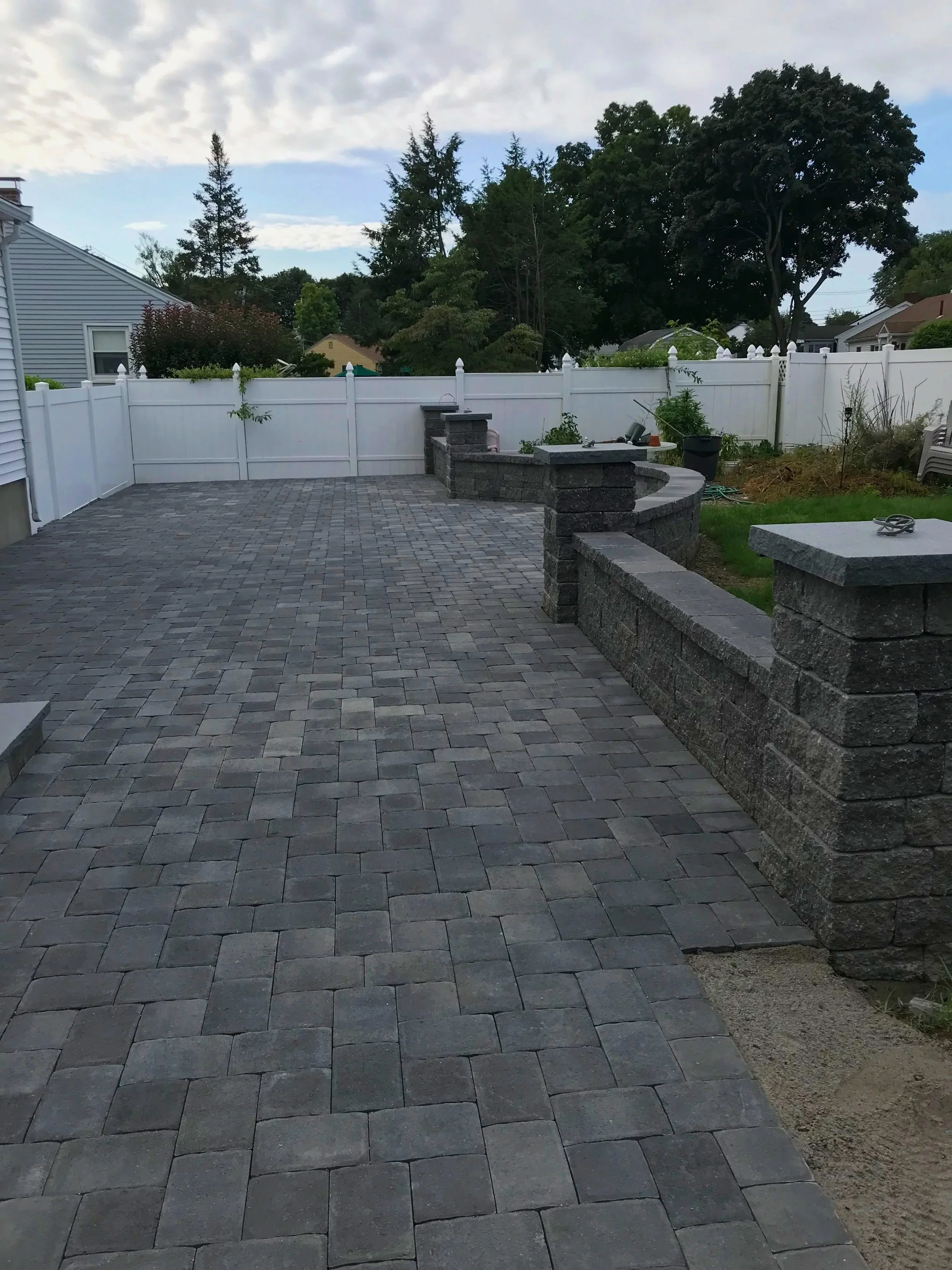 Paver patio and outdoor kitchen installation with stone retaining wall in Metrowest Boston backyard.