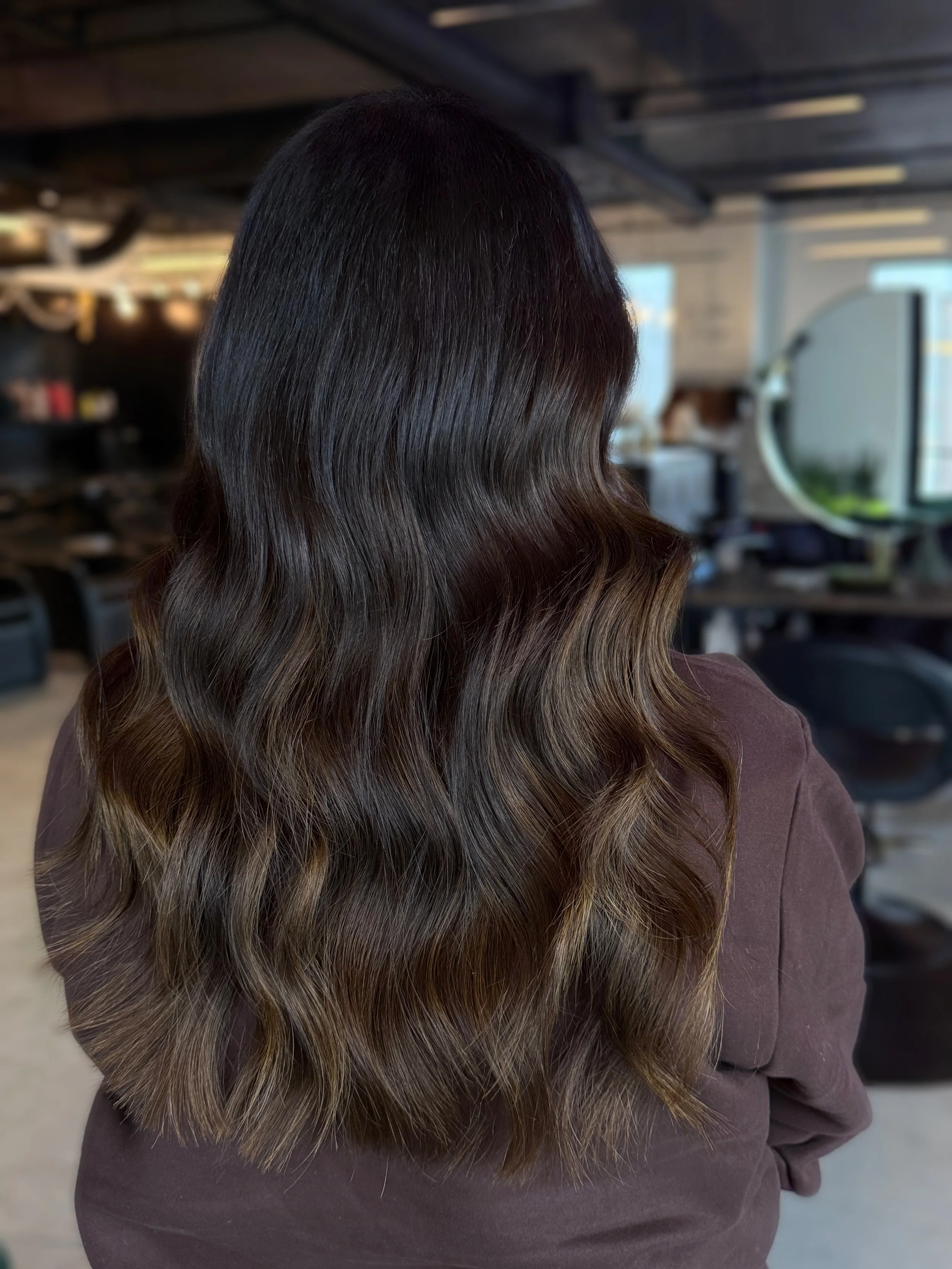 Back view of a woman with long, wavy, dark brown hair with lighter brown highlights, wearing a dark brown top, in a salon or studio setting.
