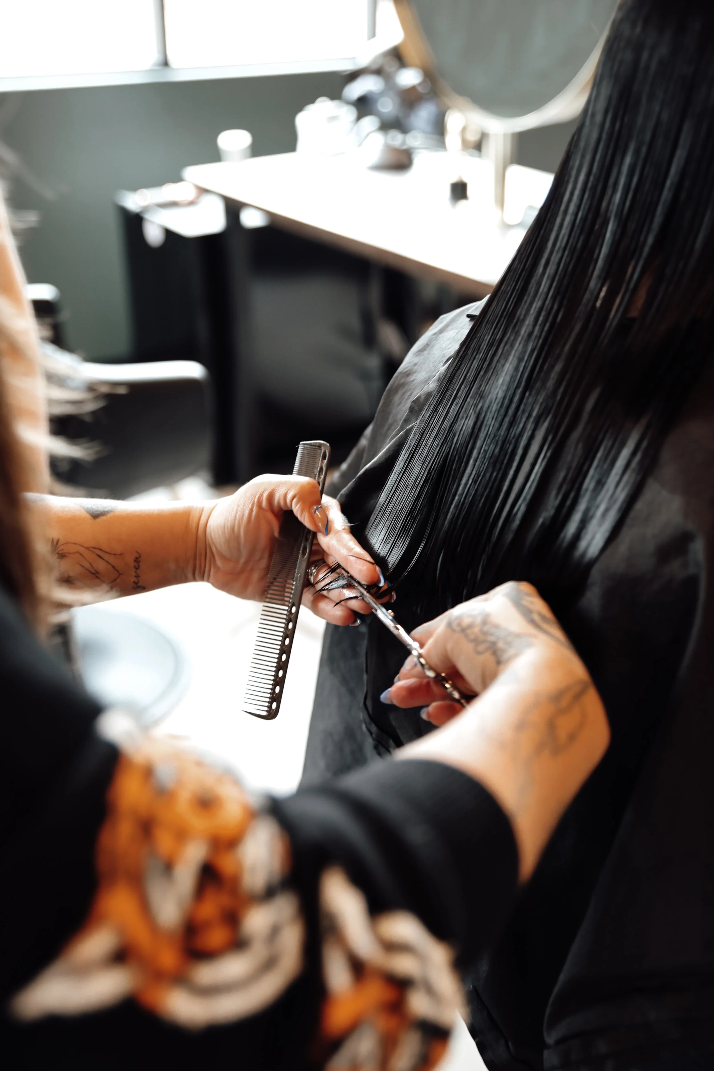 A hairstylist with tattoos is cutting a client's hair with scissors in a salon. The stylist is holding a black comb and is cutting hair in front of a mirror. The background shows salon chairs, a window, and some salon tools.