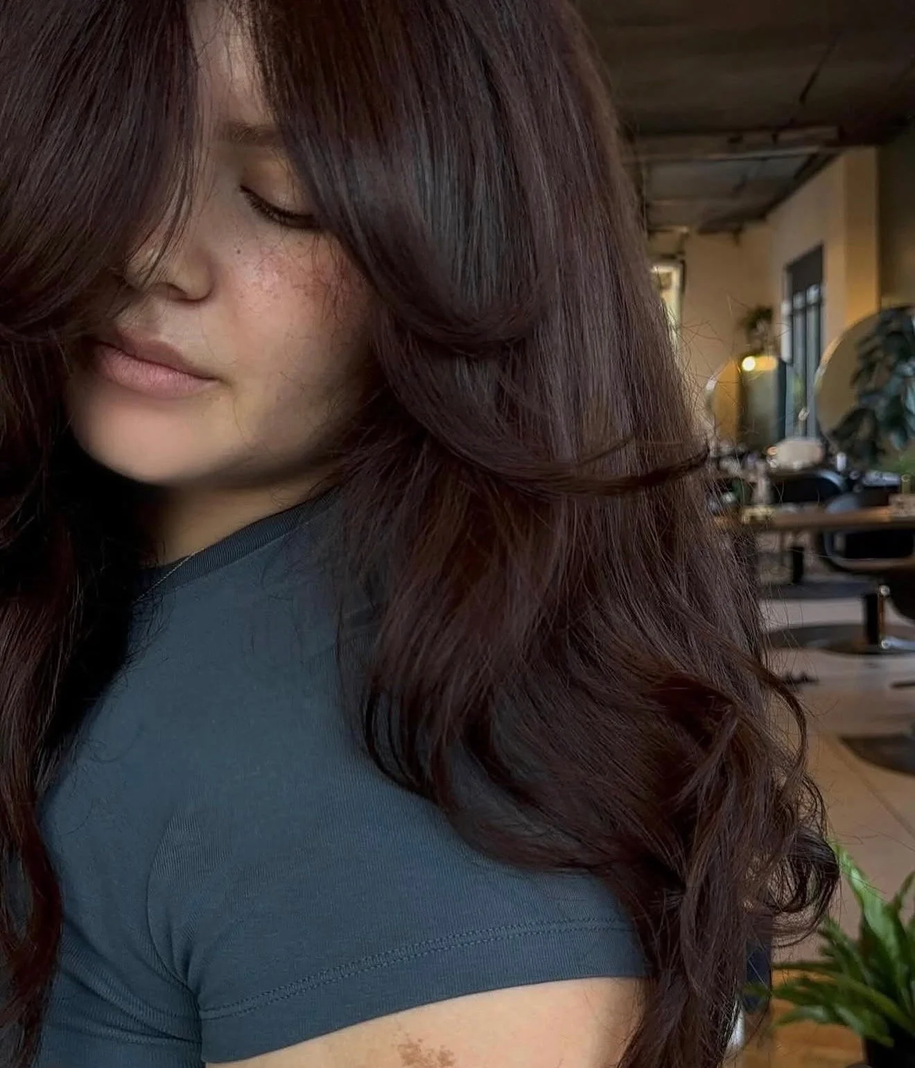 A young woman with long, wavy, dark red hair, her face partially visible with closed eyes, in a cozy indoor setting with wooden ceiling beams, plants, and furniture.