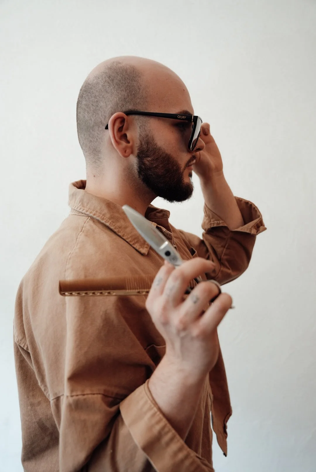 A man with a bald head and beard wearing glasses, holding a straight razor and a straight edge, adjusting his glasses with one hand, against a plain white background.