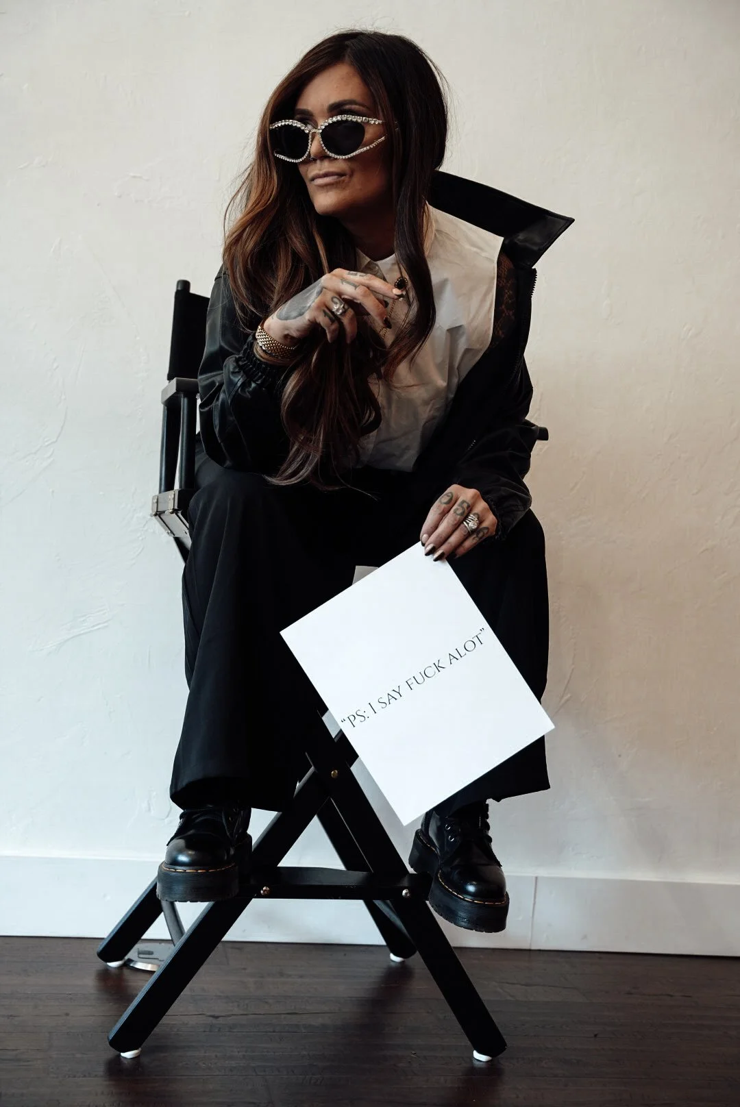 A woman with long hair and sunglasses seated on a director's chair, holding a sign that says, "P.S. I SAY F**K A LOT".