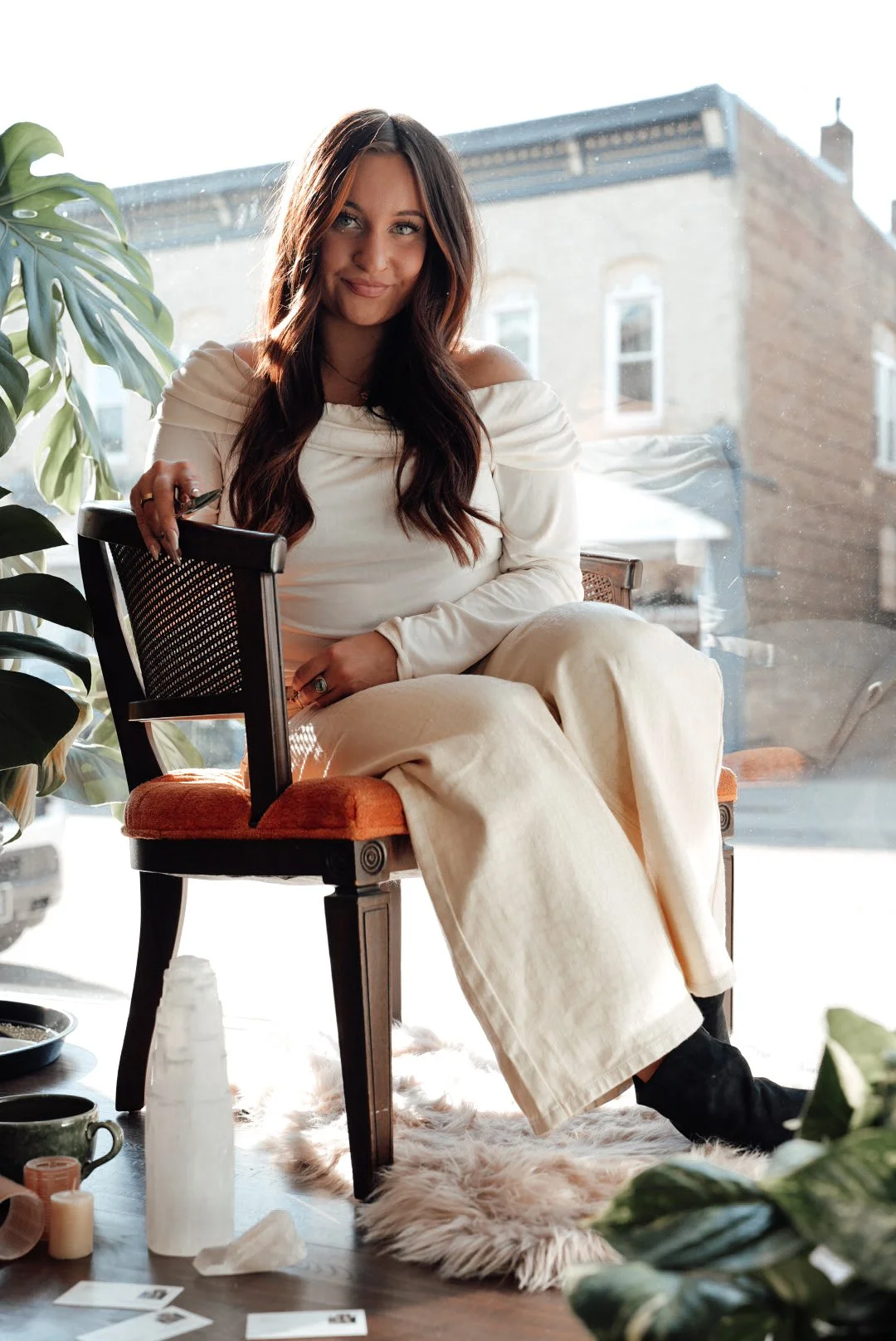 A woman with long, wavy brown hair and a warm smile sitting on an orange-cushioned chair by a large window with sunlight streaming in. She is wearing a cream-colored off-shoulder top and matching wide-legged pants, with a large green plant nearby and various small objects on a table in the foreground.
