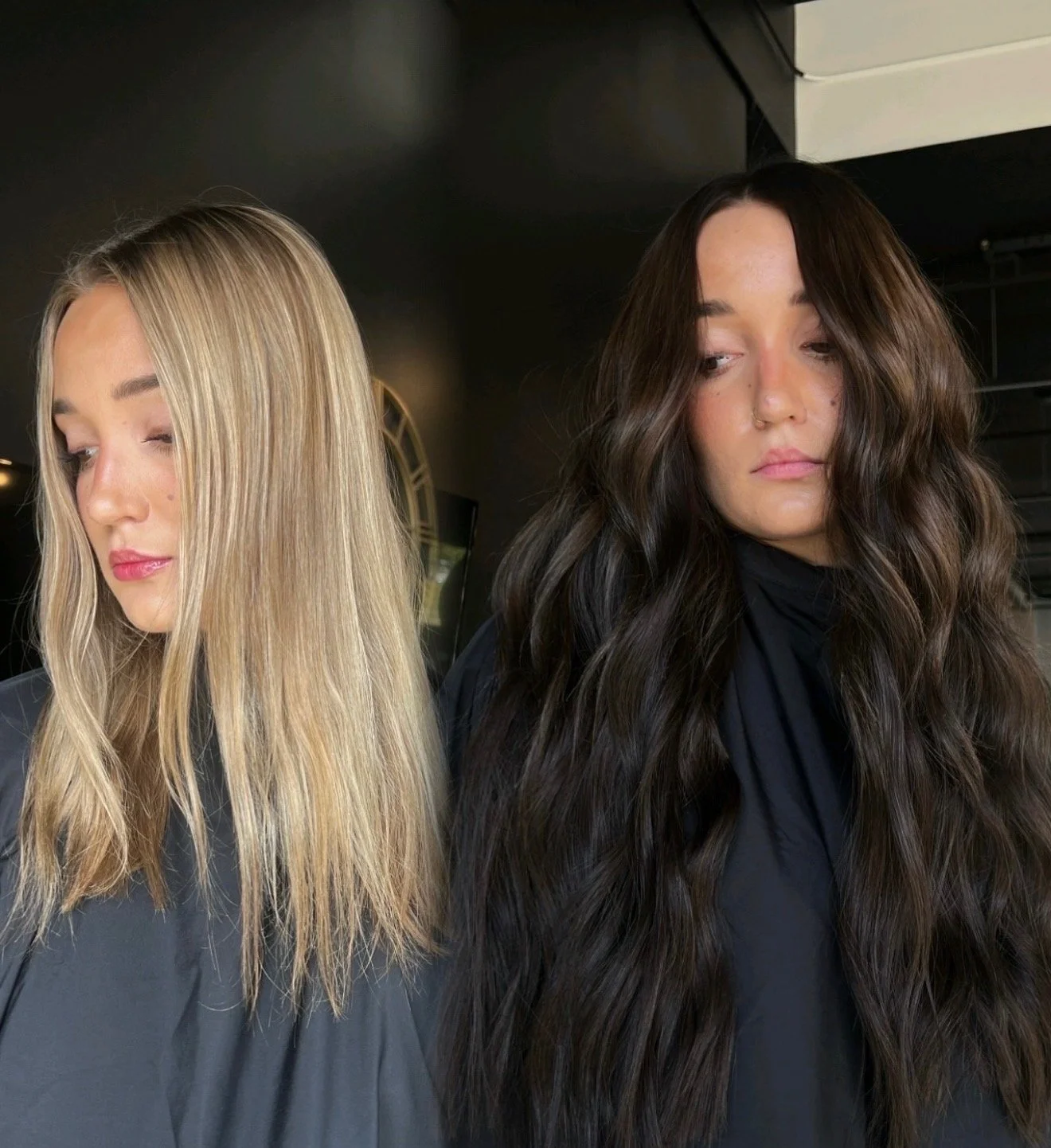 Two women with different hair colors and styles, standing side by side in what appears to be a salon, both wearing black capes. The woman on the left has long, straight, blonde hair, and the woman on the right has long, wavy, dark brown hair.