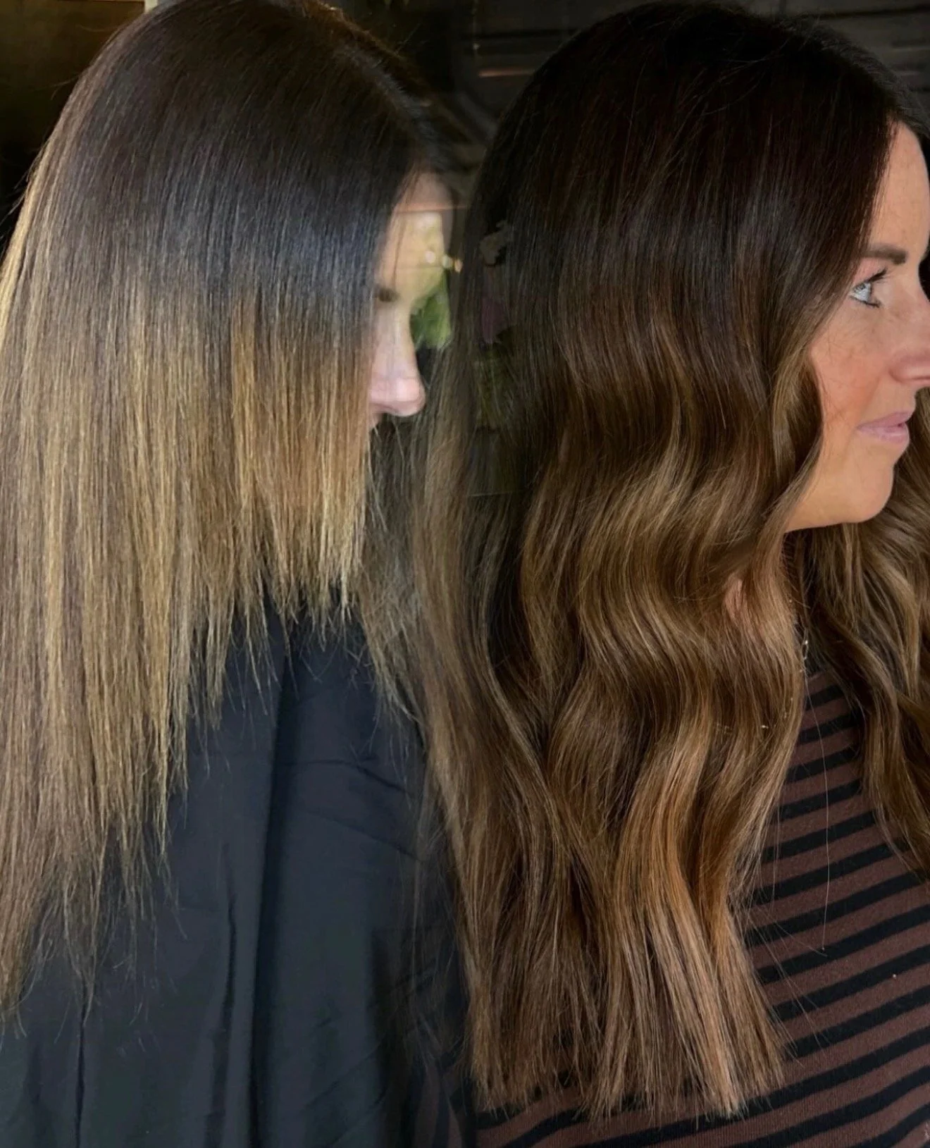 Two women with long, wavy hair, one with light brown hair and the other with dark brown hair, standing side by side indoors.